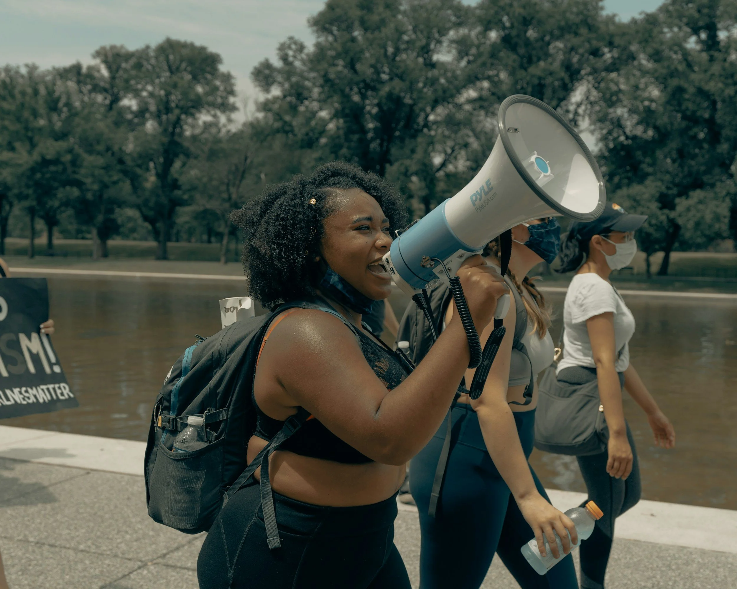 A woman with curly black hair speaking into a megaphone during a protest or rally, with other people wearing face masks walking beside her, trees and a park in the background.