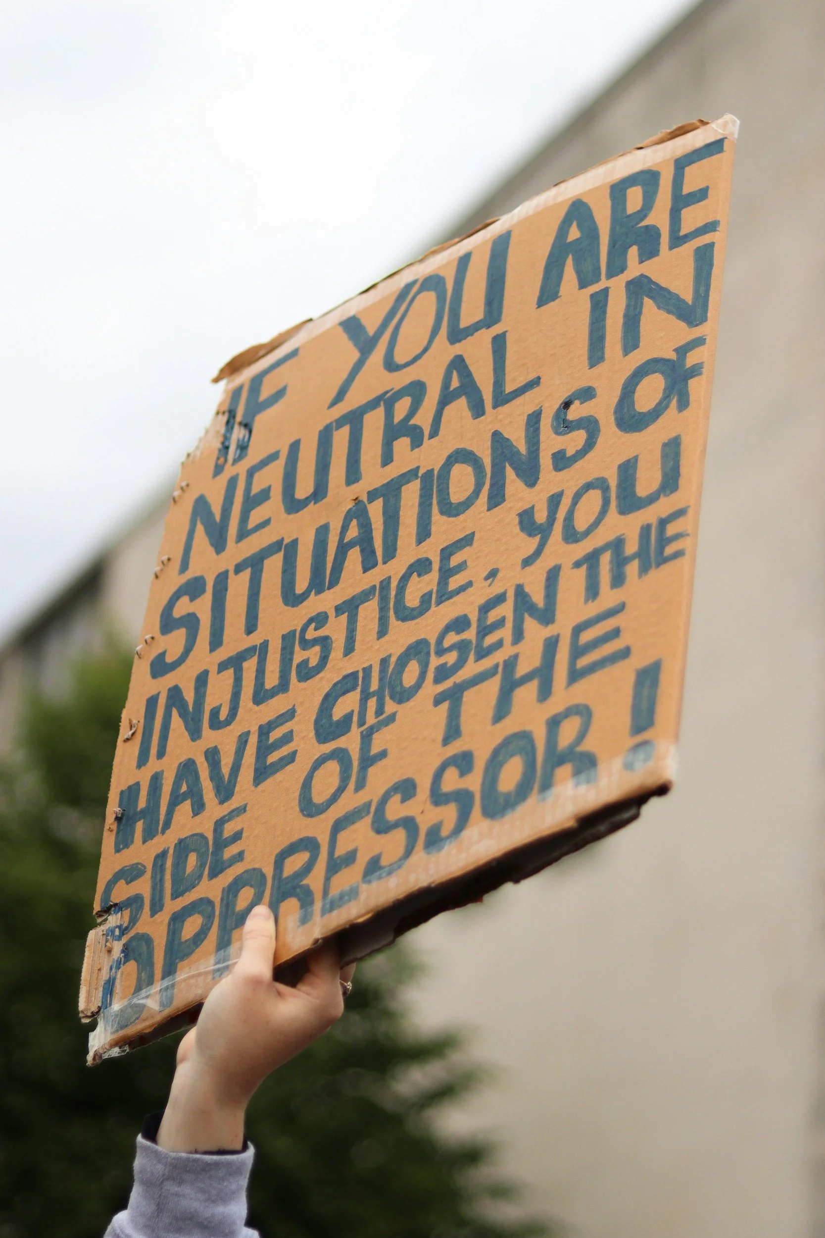 Person holding a cardboard sign with blue handwritten text at an outdoor demonstration or rally, with trees and an overcast sky in the background.