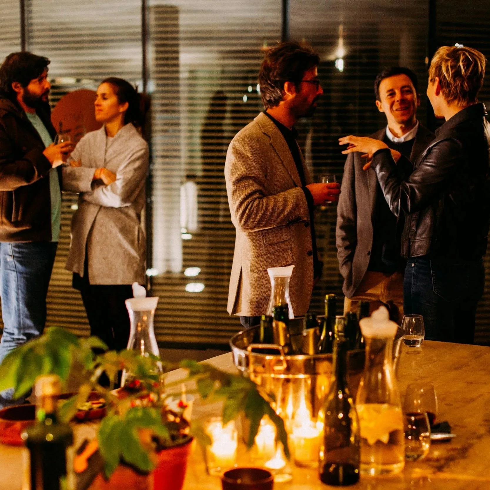 Group of people talking and socializing at a dinner party or gathering in an indoor setting with warm lighting, glasses, and a table with bottles and decor.