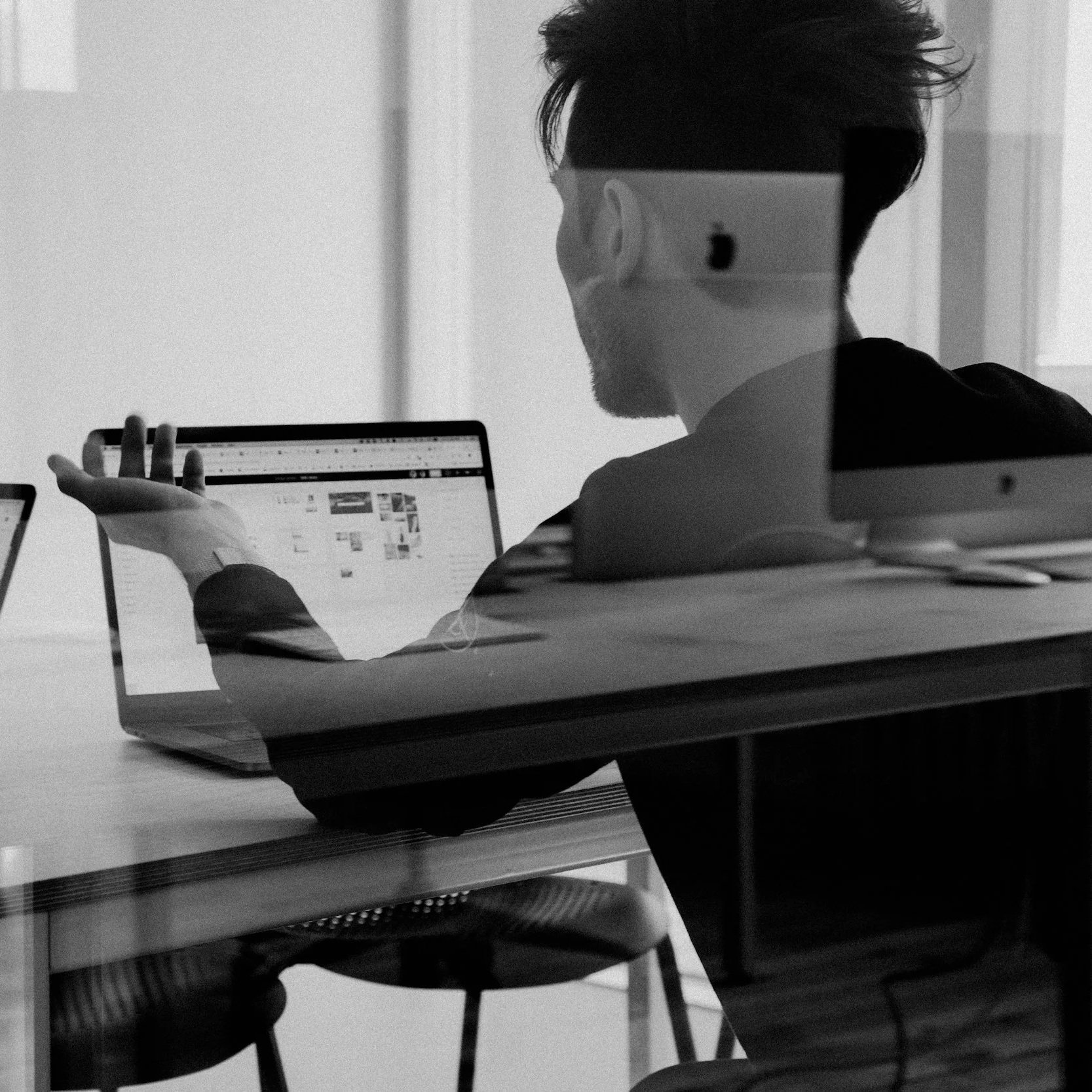 Black and white photo of a man sitting at a desk and using a tablet in a modern office. He is wearing a face mask and a hoodie, with messy hair. The photo is taken through a glass window, reflecting light.