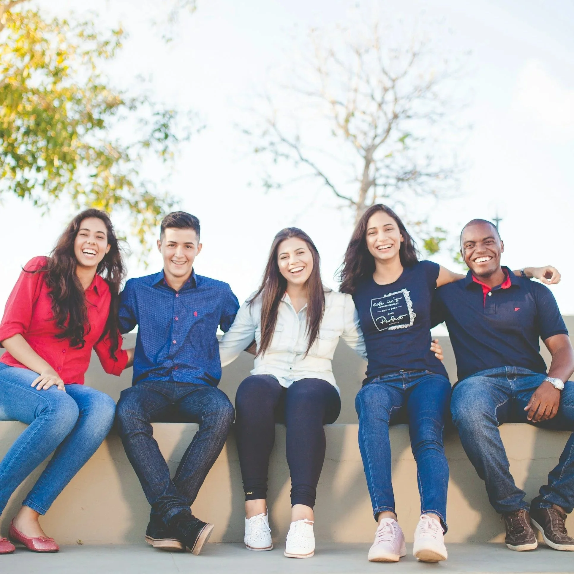 Group of diverse young adults sitting on a ledge outdoors, smiling and laughing, with a tree and blue sky in the background.