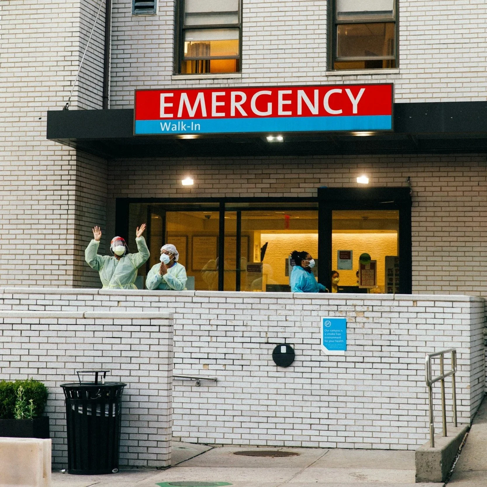 Hospital emergency walk-in entrance with three healthcare workers wearing masks and personal protective equipment, standing outside on the sidewalk.