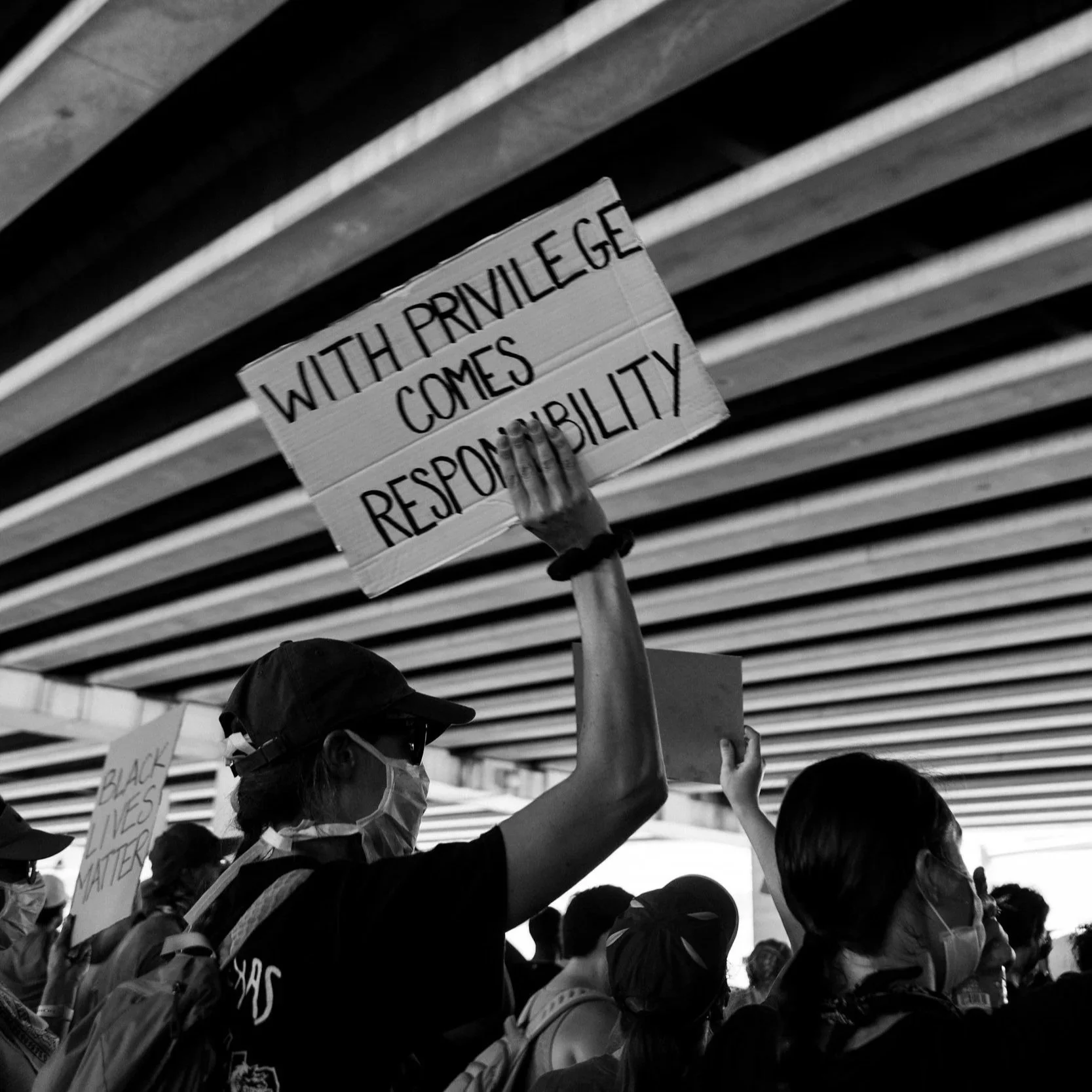 Protesters wearing face masks under a bridge, holding signs including one that reads 'With Privilege Comes Responsibility' in black and white.