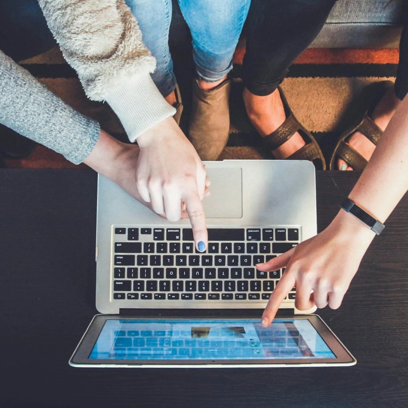 Two people are using a laptop and a tablet on a table, both pointing at the laptop keyboard.