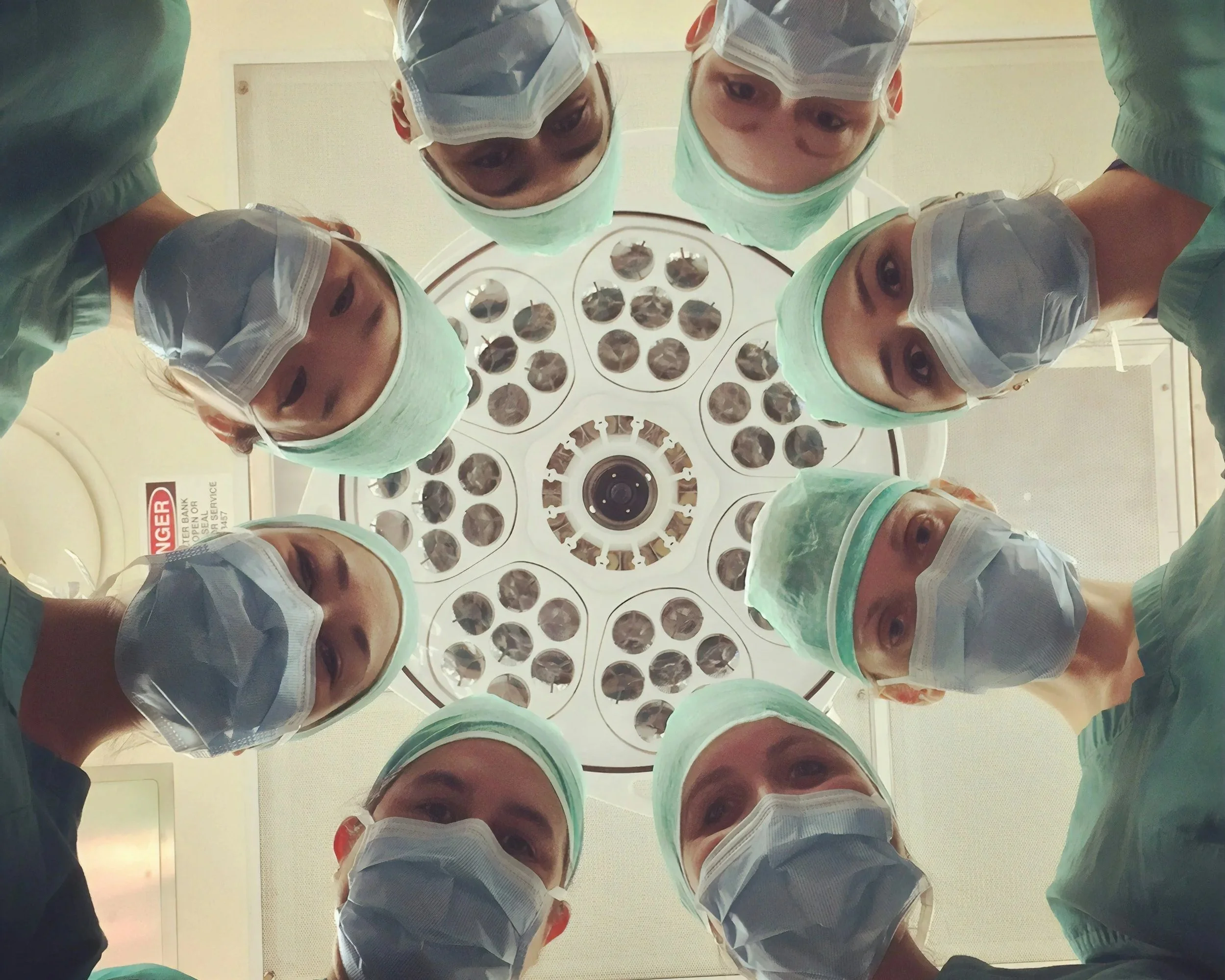 A group of surgeons in scrubs, masks, and caps leaning over an operating room light, looking down into the camera.