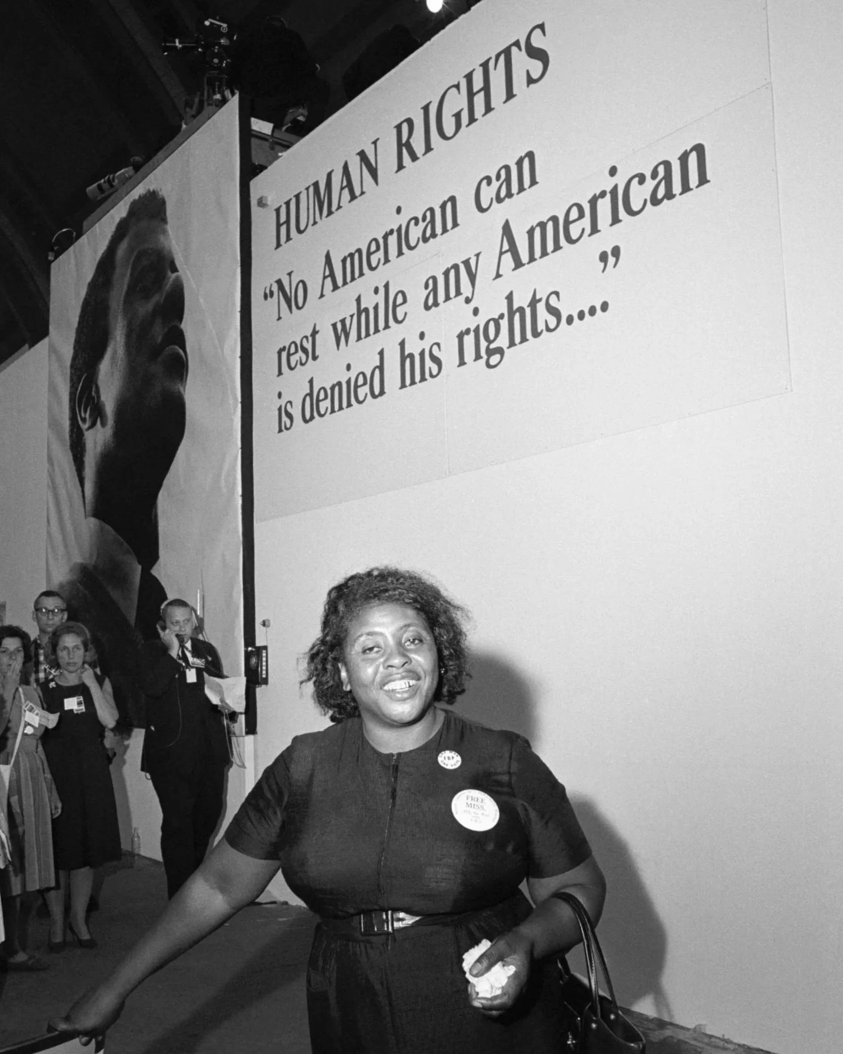 Black and white photo of a woman with a bandaged finger smiling at the camera, wearing a dark dress with buttons and a sign that says 'Free Miss.' behind her, with people in the background and a large sign that reads 'Human Rights No American Can Rest While Any American is Denied His Rights.'