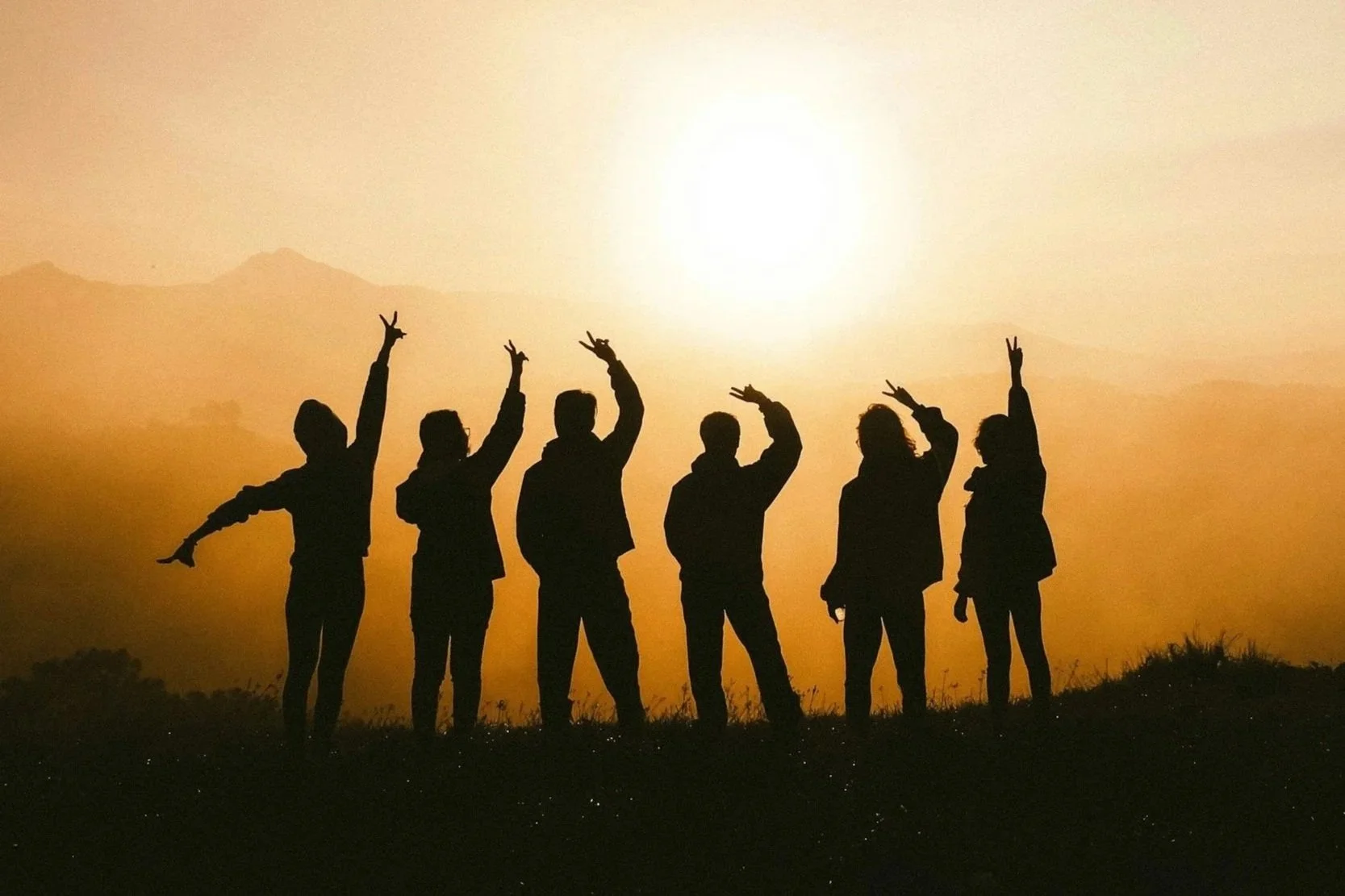 Six silhouetted people standing on a grassy hill during sunset, making peace signs with their hands against a bright setting sun and distant mountains.