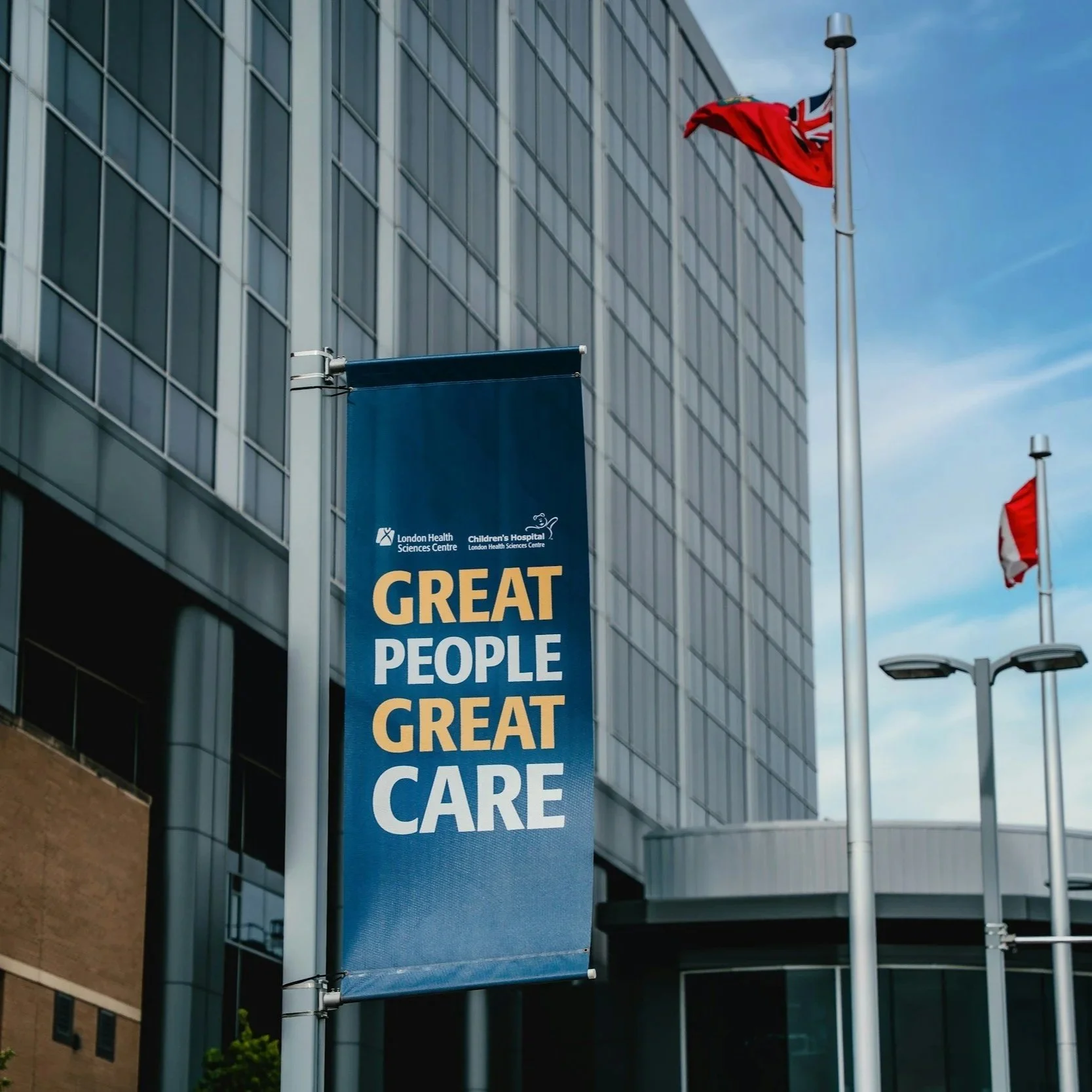 Blue banner with white and yellow text on a building reads, 'GREAT PEOPLE GREAT CARE.' Two red flags with Union Jack on pole, modern office building in background.