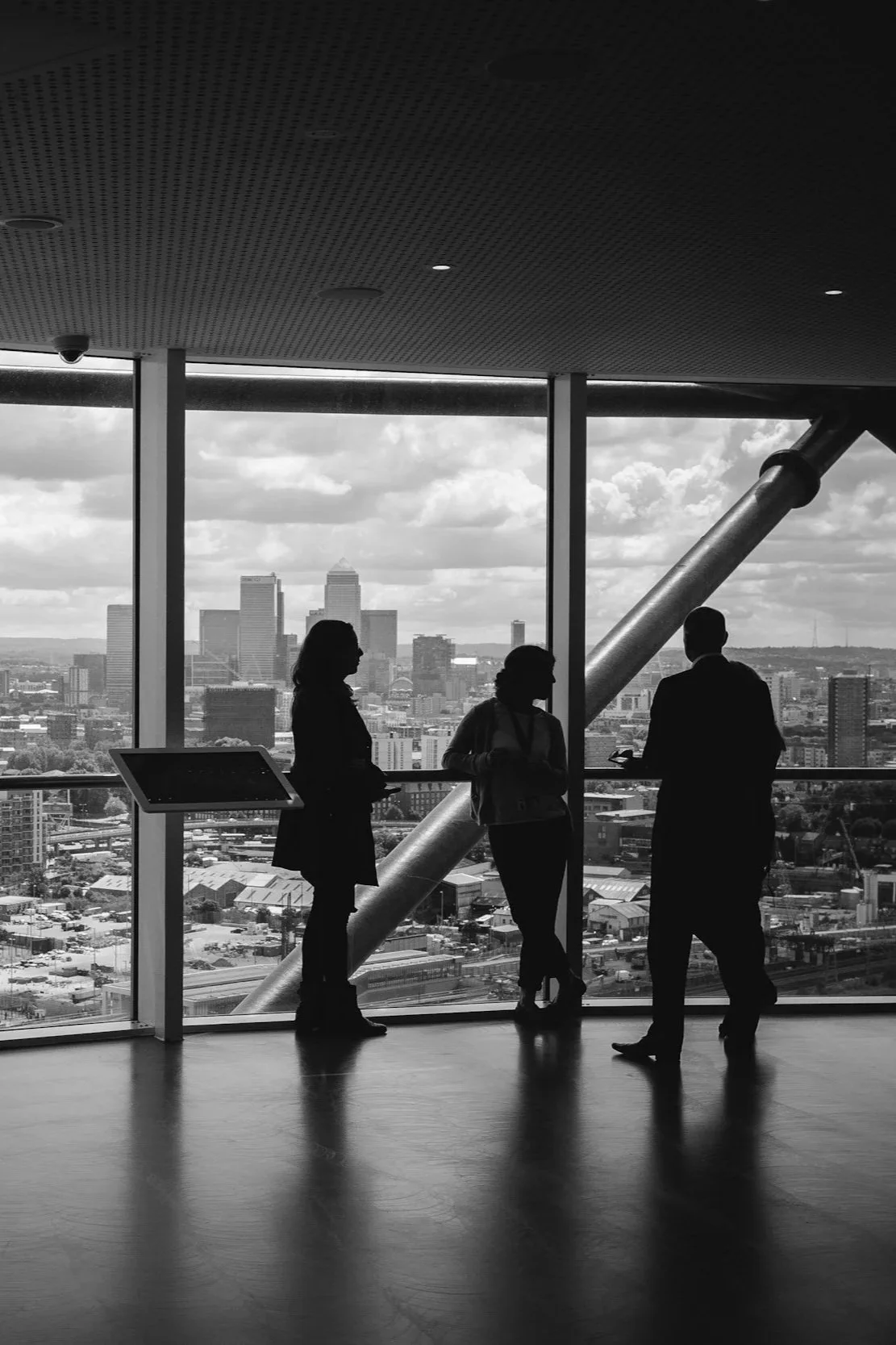 Silhouettes of three people talking inside a high-rise building with a city skyline view in the background that includes tall buildings and a cloudy sky.