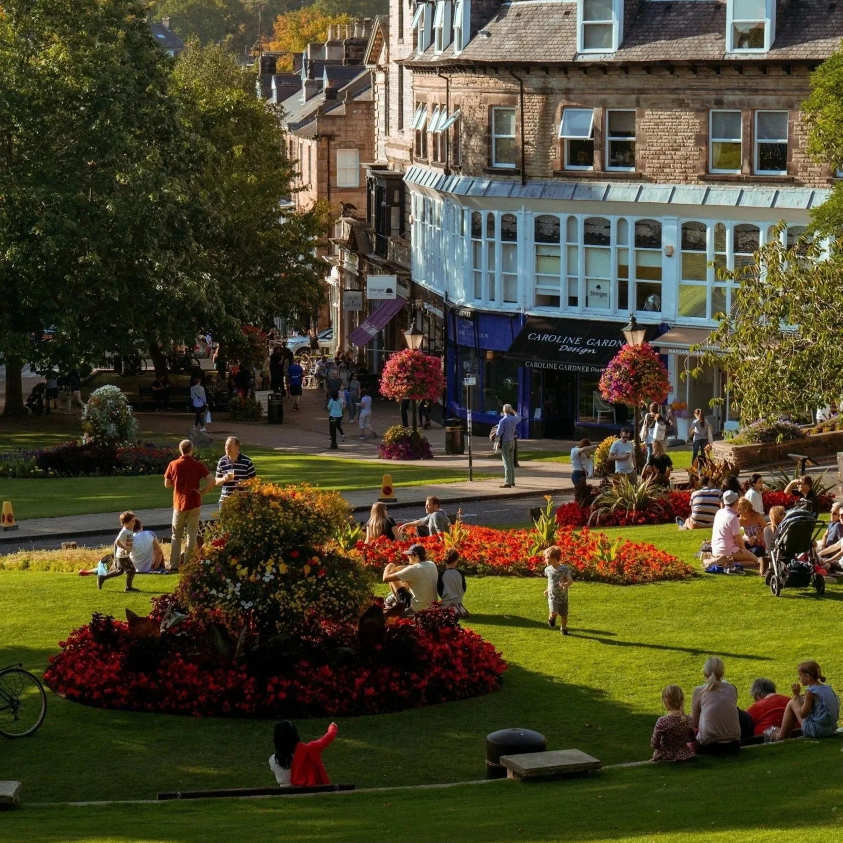 A lively town square with people sitting and walking, surrounded by lush greenery and colorful flower beds, with historic brick buildings in the background.