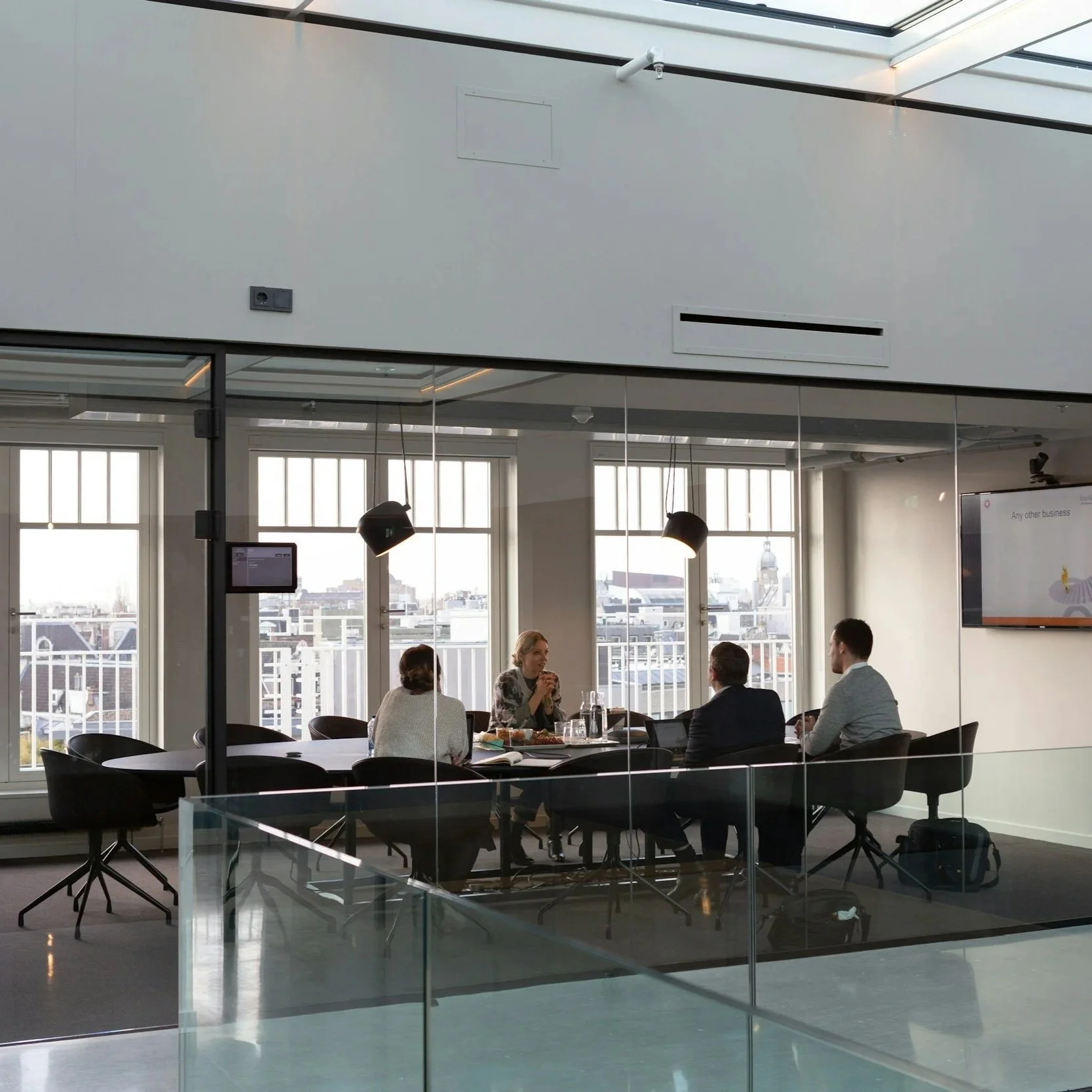 A group of four people sitting around a modern round conference table in a glass-walled office with large windows, discussing and engaging in conversation.
