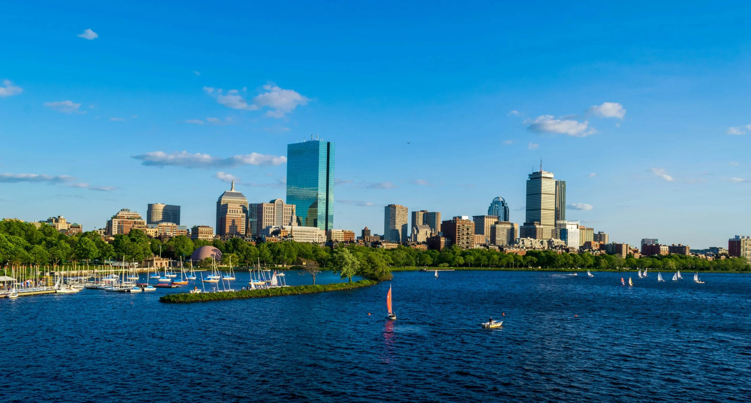 Boston skyline view with skyscrapers, a body of water in the foreground, trees, and sailboats under a blue sky with scattered clouds.