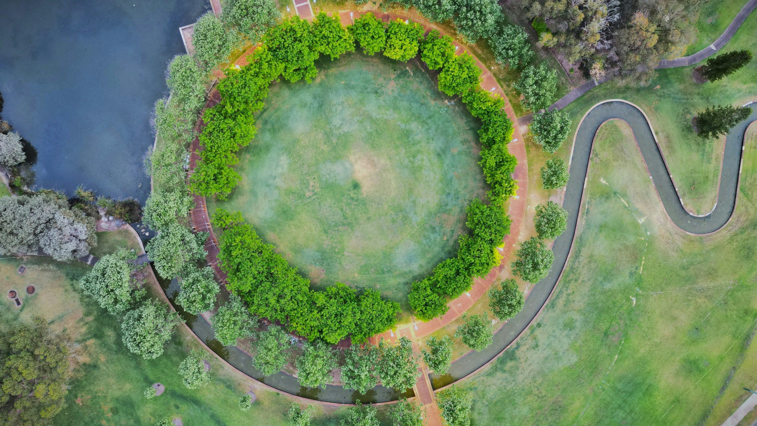 aerial view of a circular park with a grassy center surrounded by a pathway, lined with green trees, near a blue body of water with a small bridge