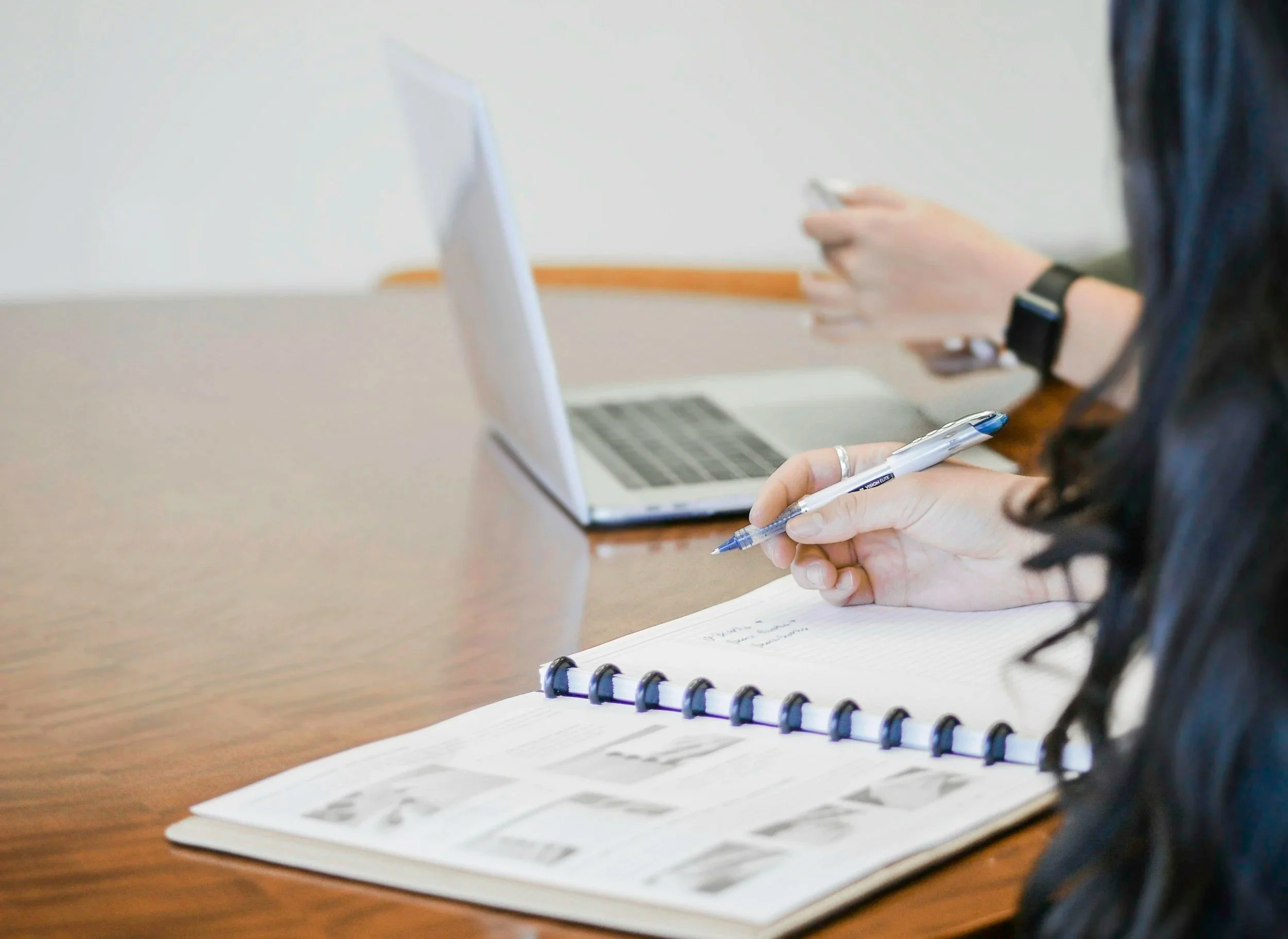 Person writing in a notebook with photos, sitting at a wooden table, with a laptop and another person using a smartphone in the background.