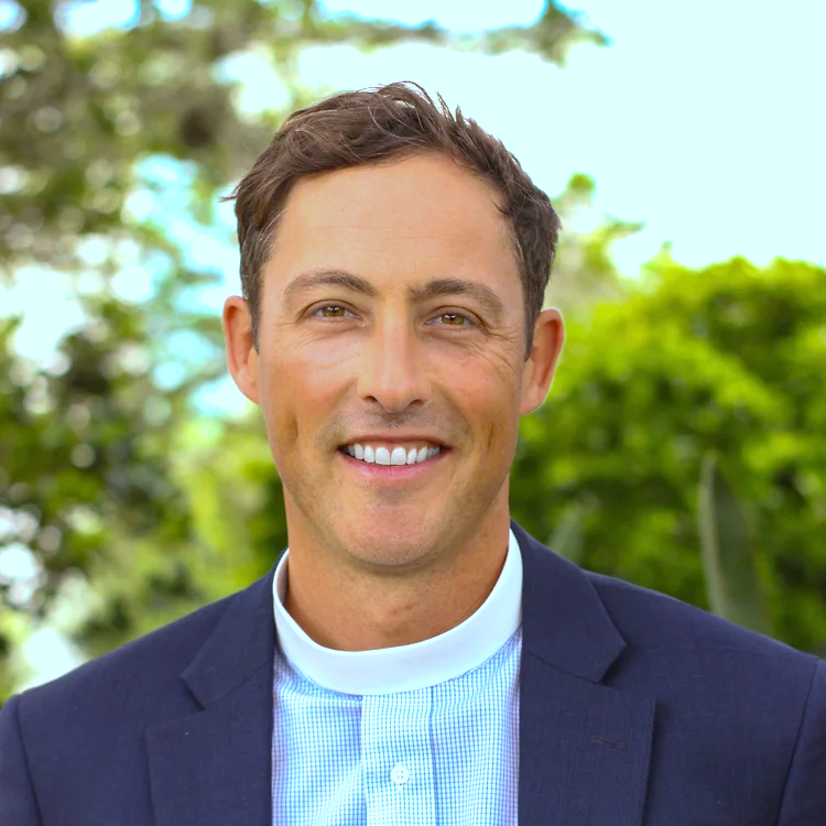 A man smiling outdoors with trees and blue sky in the background.