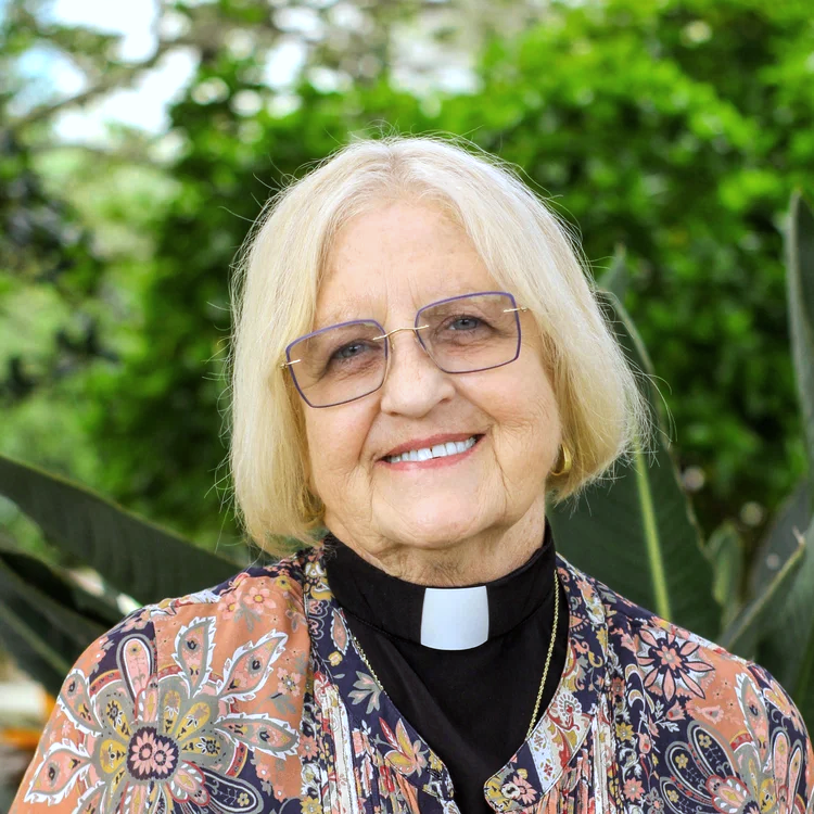 A smiling elderly woman wearing glasses and a clerical collar, standing outdoors with green foliage in the background.