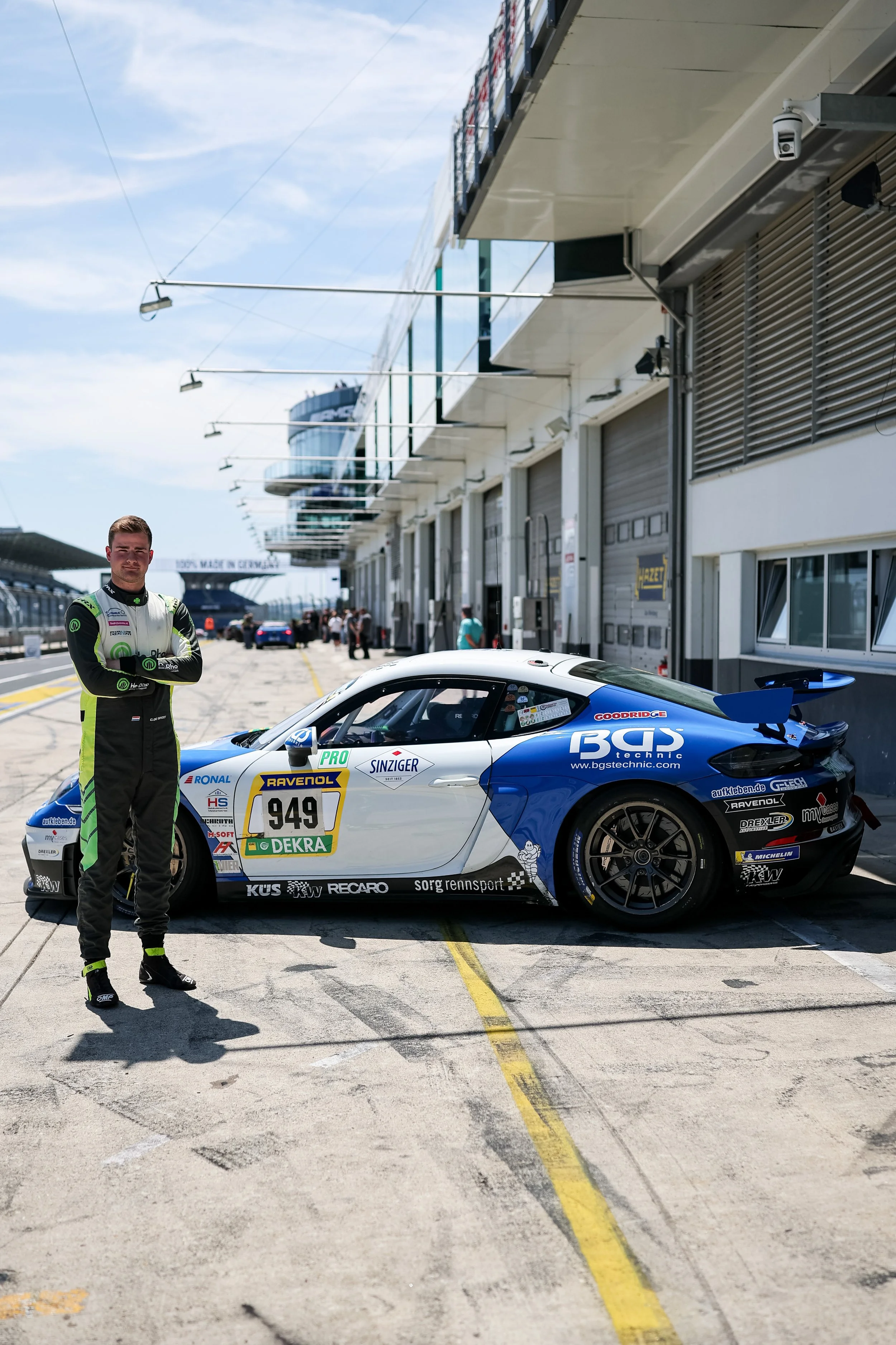A race car driver in a racing suit stands beside a race car on a racing track.