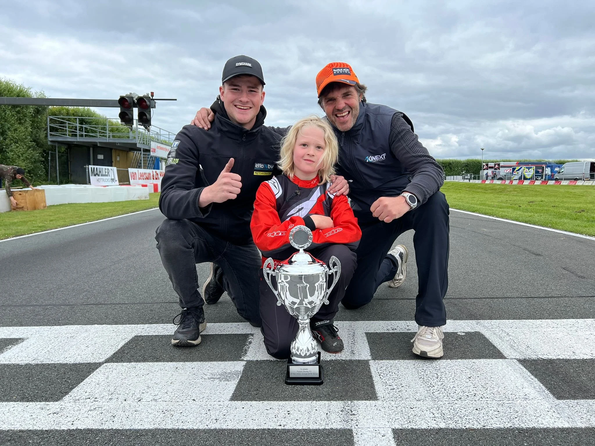 Three people kneeling on a race track with a trophy, celebrating after a race, with overcast skies and racing trailers in the background.
