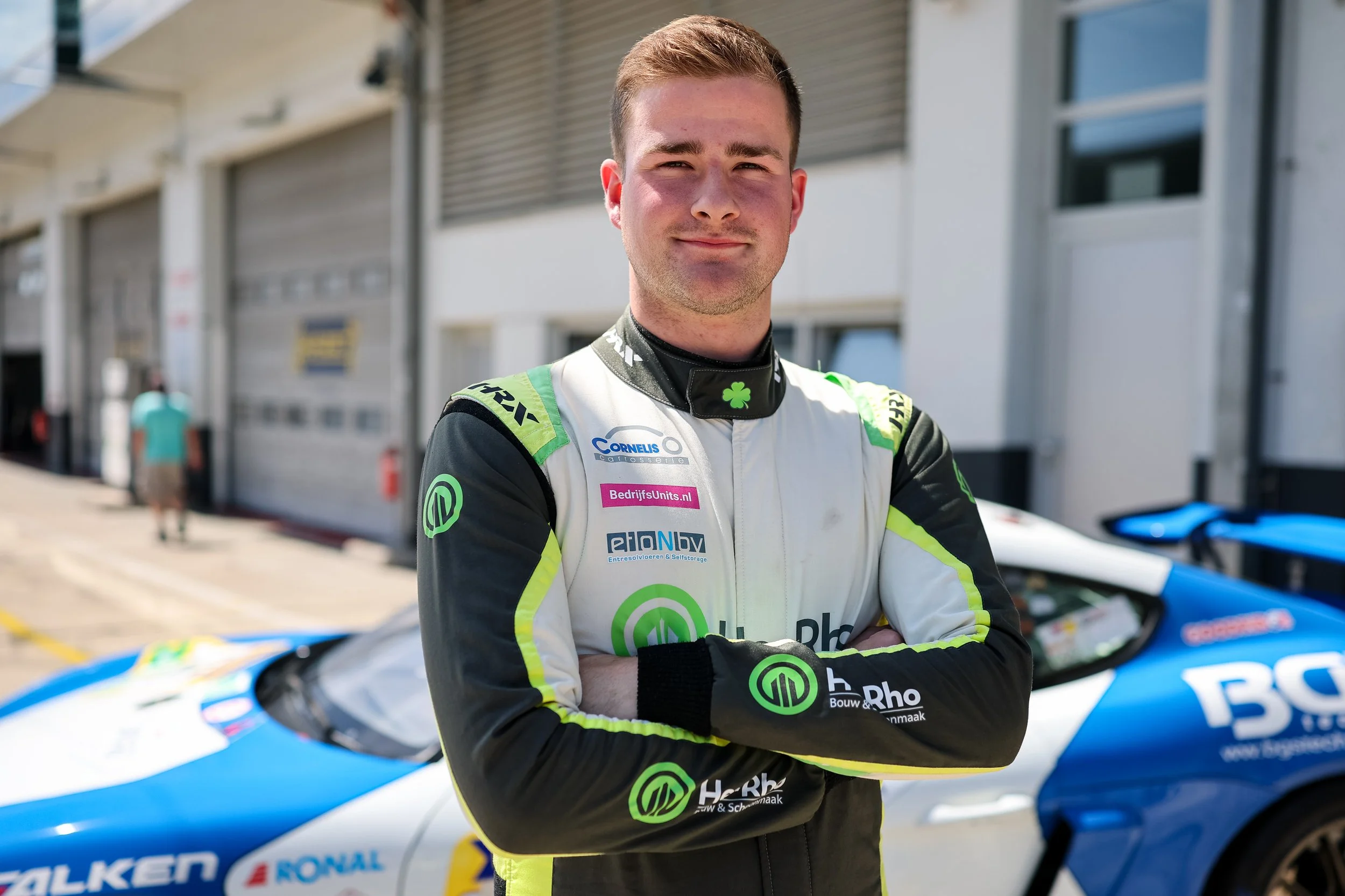 A male race car driver in a racing suit standing with arms crossed in front of a blue and white race car, at a racetrack with garage doors in the background.