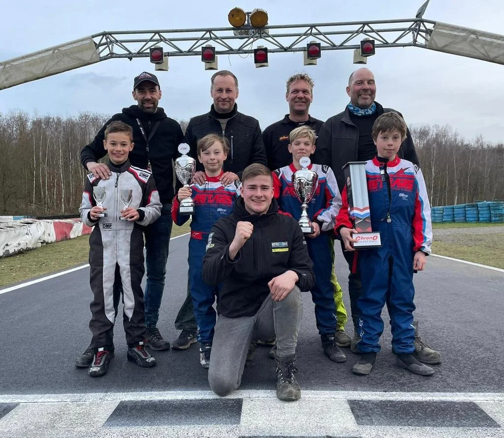 Group of seven people, including five children and two adults, standing on a race track holding trophies after a race event, with race barriers and trees in the background.