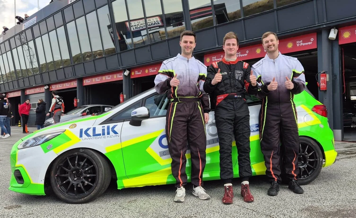 Three race car drivers standing in front of a race car with a green and white livery at a racing event, with a pit building and other people in racing suits in the background.