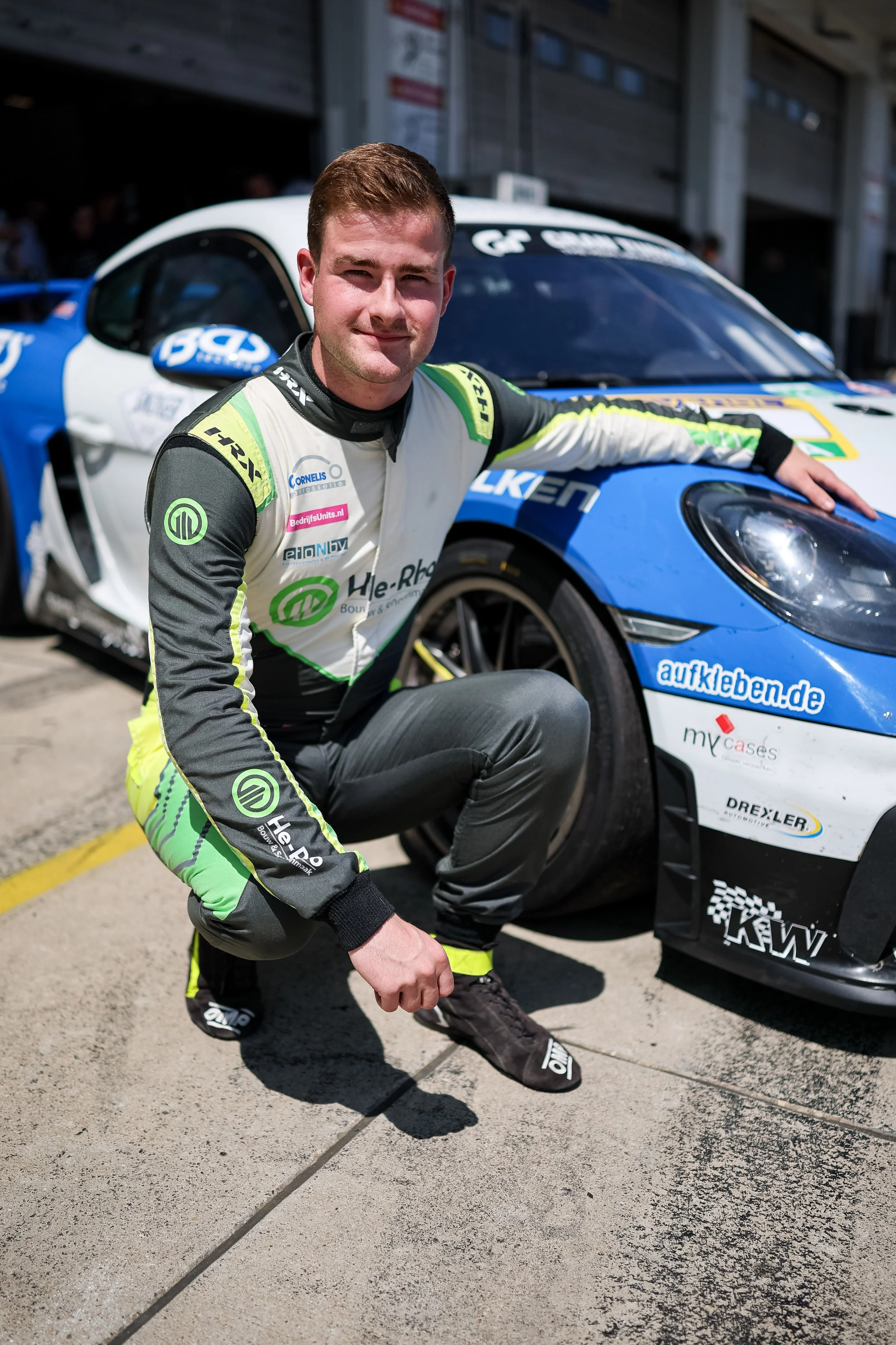 A race car driver squatting beside his blue and white race car, wearing a racing suit with sponsor logos, in a pit lane area.