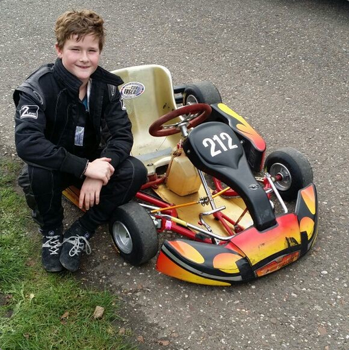 A young boy with a big smile posing next to a small go-kart with the number 212, parked on a paved surface with grass nearby.