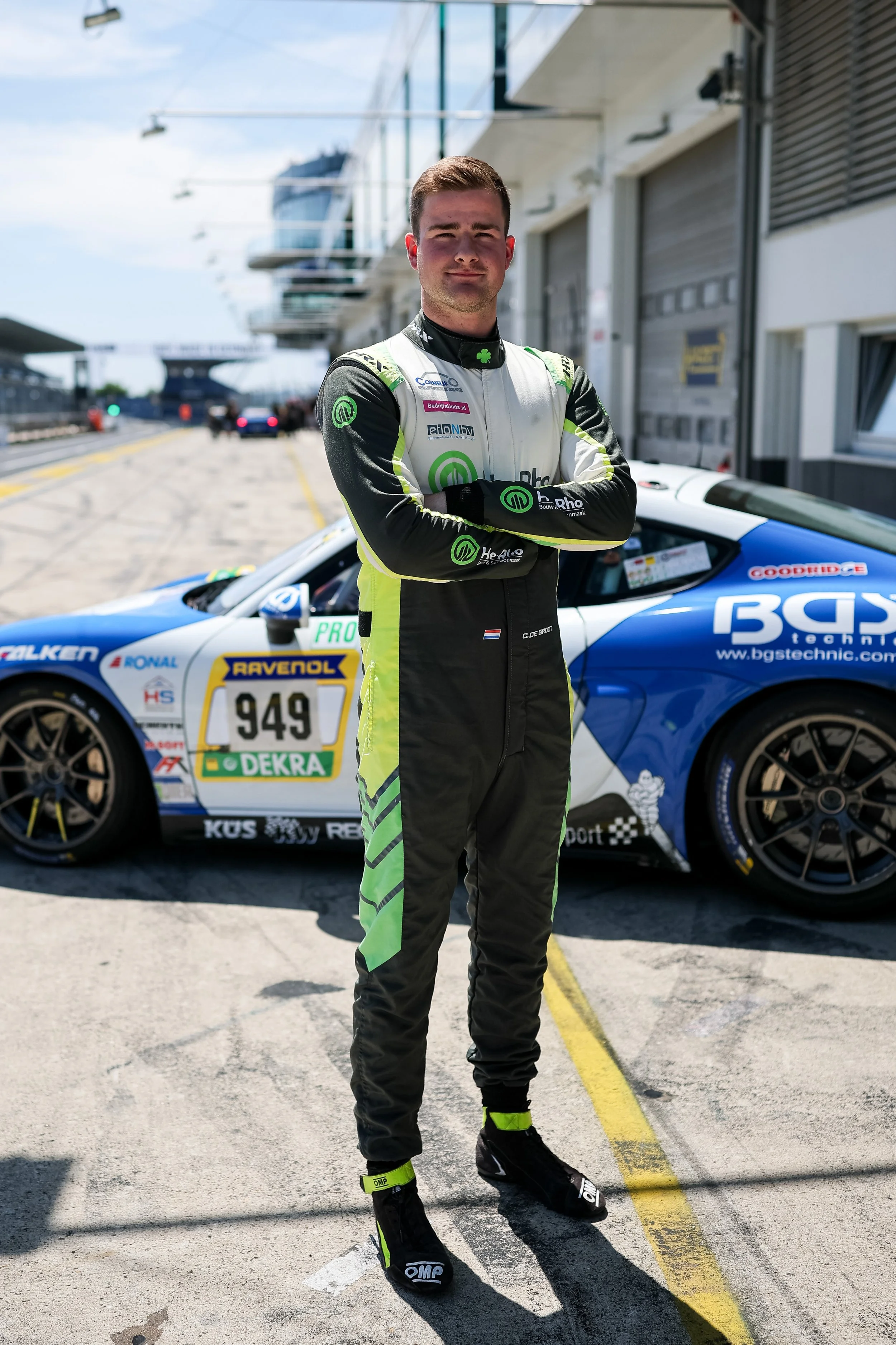 A male race car driver in a racing suit with crossed arms, standing in front of a blue and white race car on a pit lane.