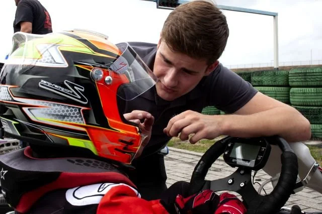 A young man leaning over a go-kart steering wheel, talking to a person wearing a colorful racing helmet.