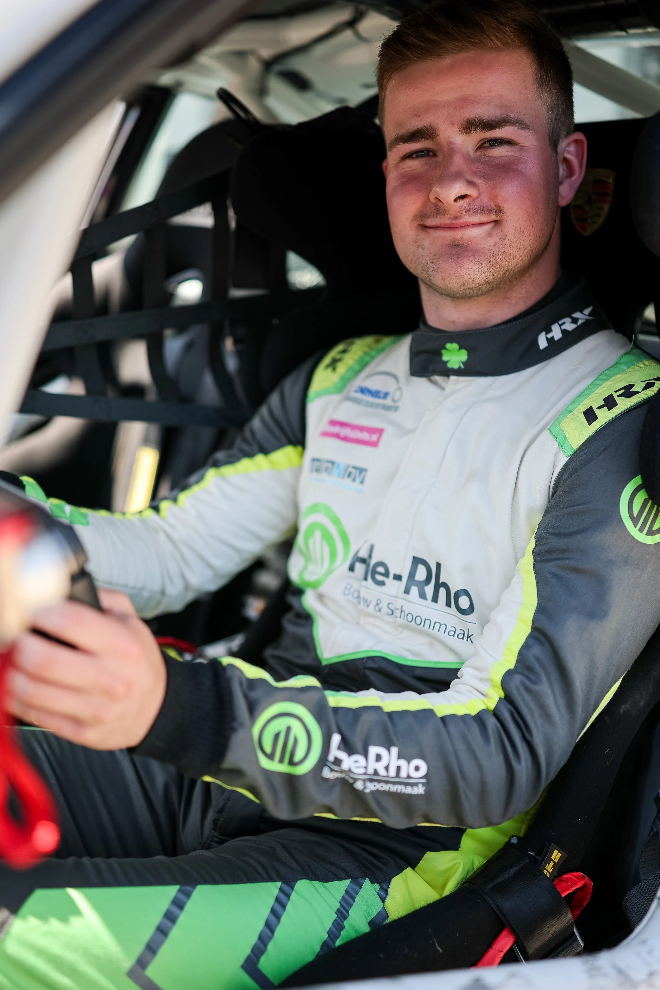 A race car driver in a racing suit sitting inside a car, smiling at the camera. The suit features various sponsor logos including He-Rho, I-R18, and others. The driver is seated in a racing seat with a harness and a safety cage visible behind him.