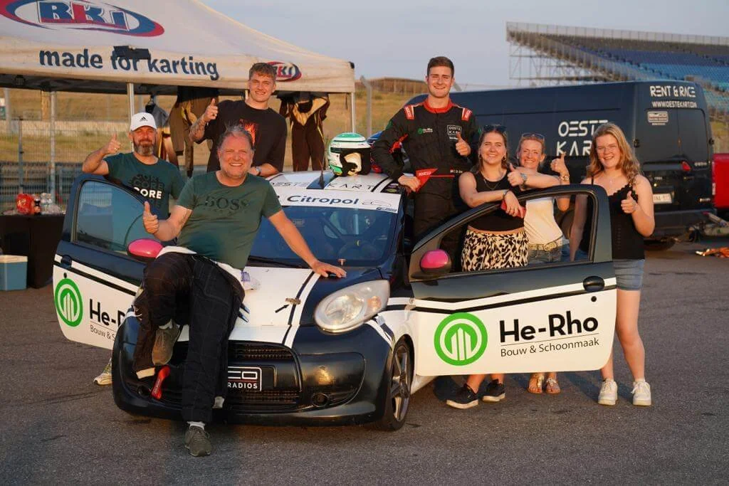 Group of people celebrating around a small racing car with a helmet on top, outdoors at a racetrack with a tent and a truck in the background, all giving thumbs up.