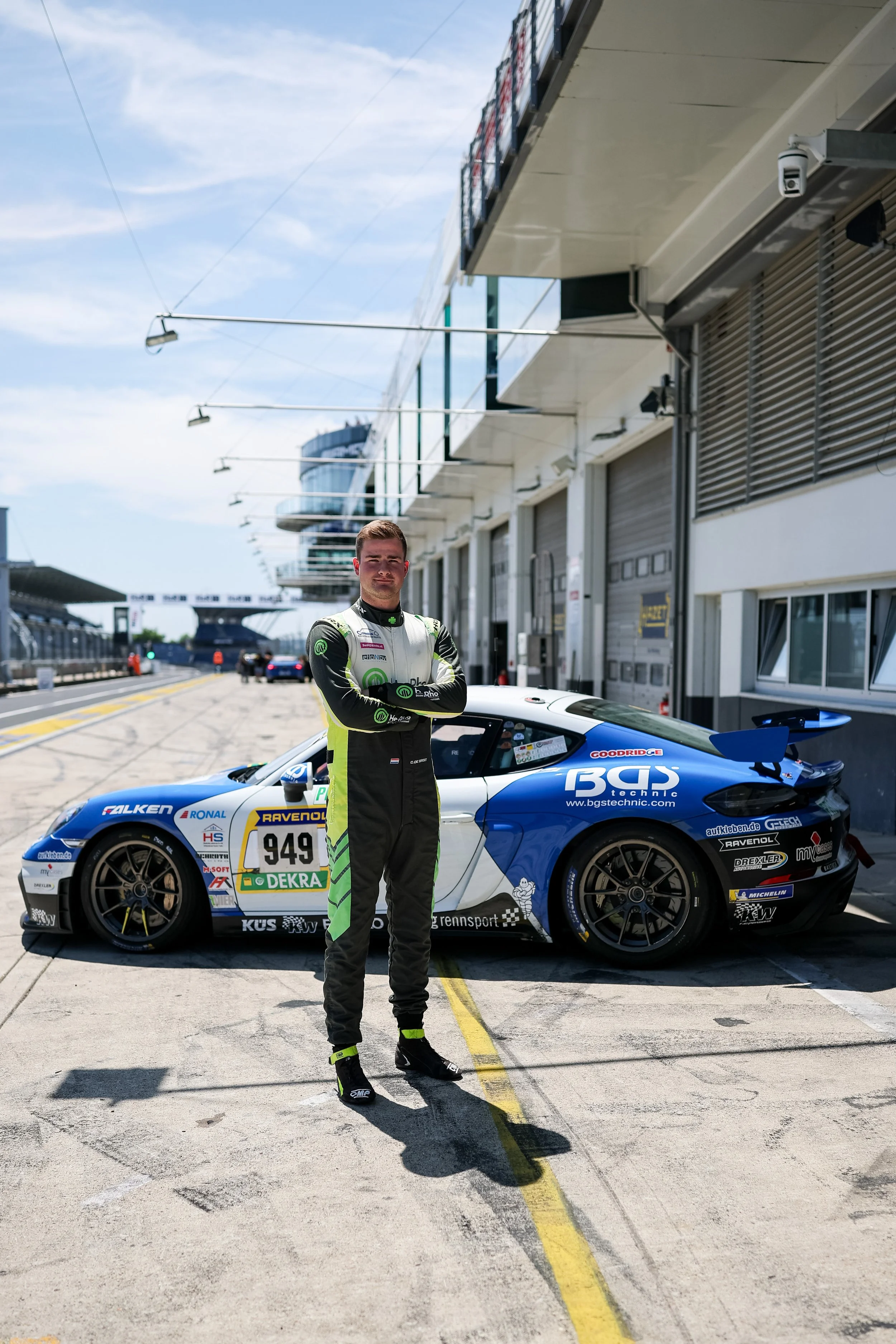 Race car driver standing with arms crossed in front of a blue and white race car at a racing circuit.