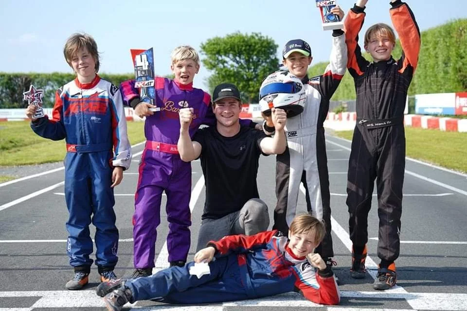 Children celebrating on a race track, holding trophies and wearing racing suits, with a smiling young man kneeling in the center.