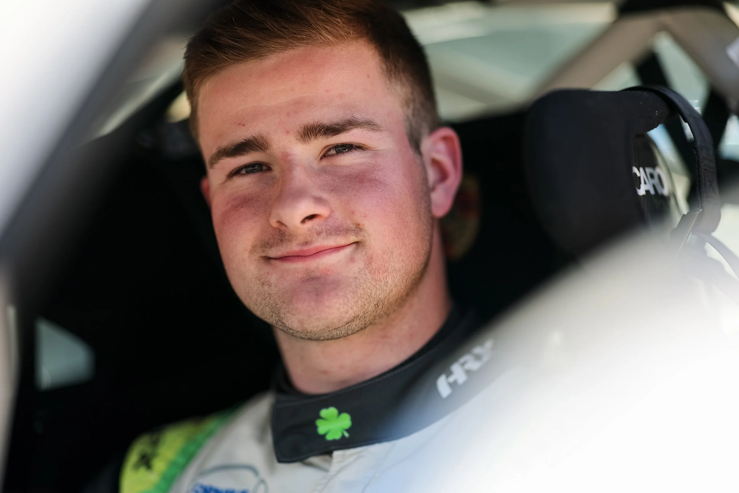 Close-up of a young man sitting in a race car, smiling and looking at the camera, wearing a racing suit with a shamrock badge on the collar.