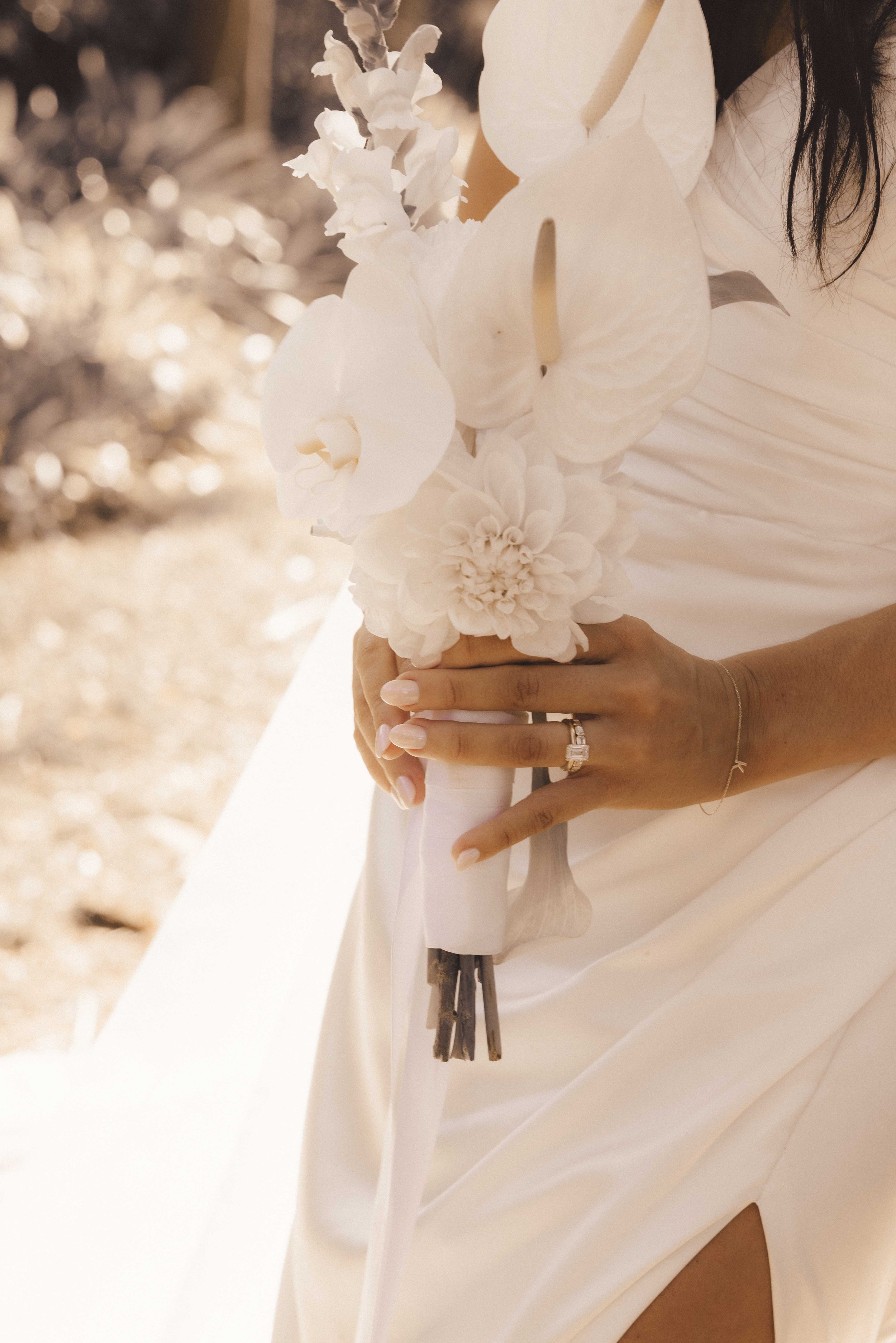 Close-up of a woman in a white dress holding a bouquet of white flowers, with her hand showing a wedding ring, on a bright background.