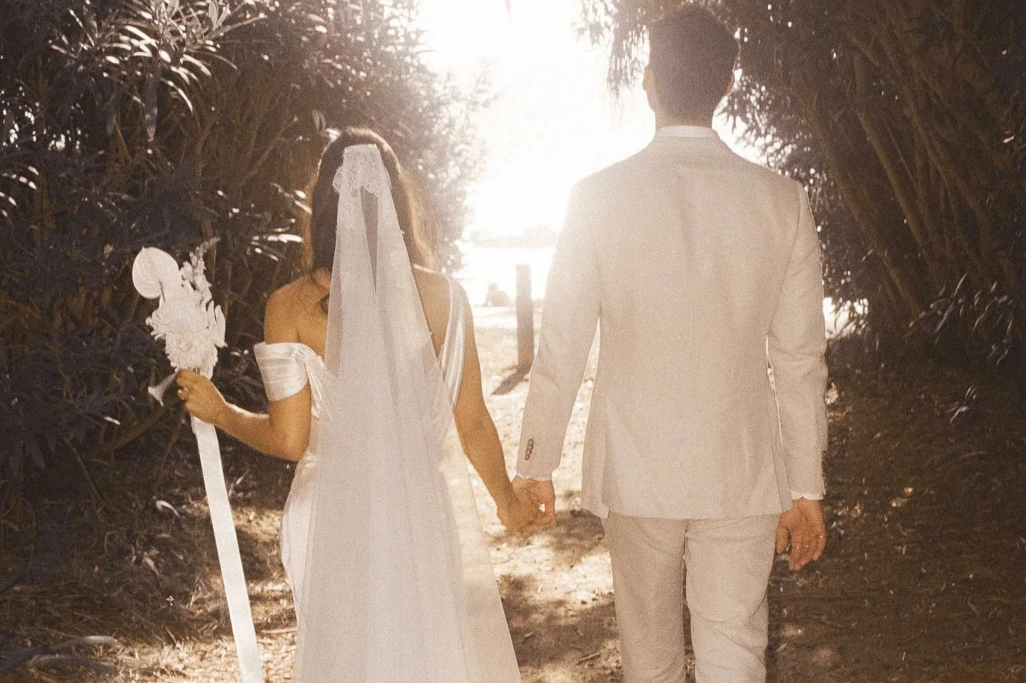 A couple dressed in wedding attire walking hand in hand through a lush, green, tree-lined pathway during sunset or sunrise.