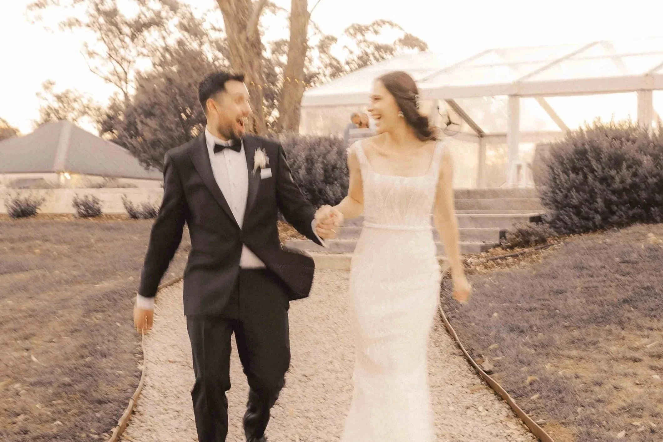 A man and woman in wedding attire holding hands and smiling while walking outdoors during sunset.