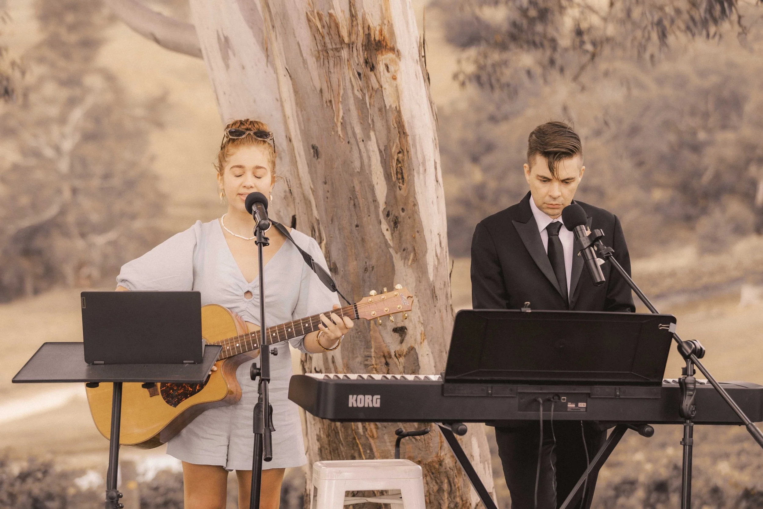 A woman with red hair playing an acoustic guitar and singing into a microphone, wearing a light-colored dress, and a man with dark hair in a black suit playing a keyboard and singing into a microphone, outdoors near a large tree, with desert-like landscape in the background.