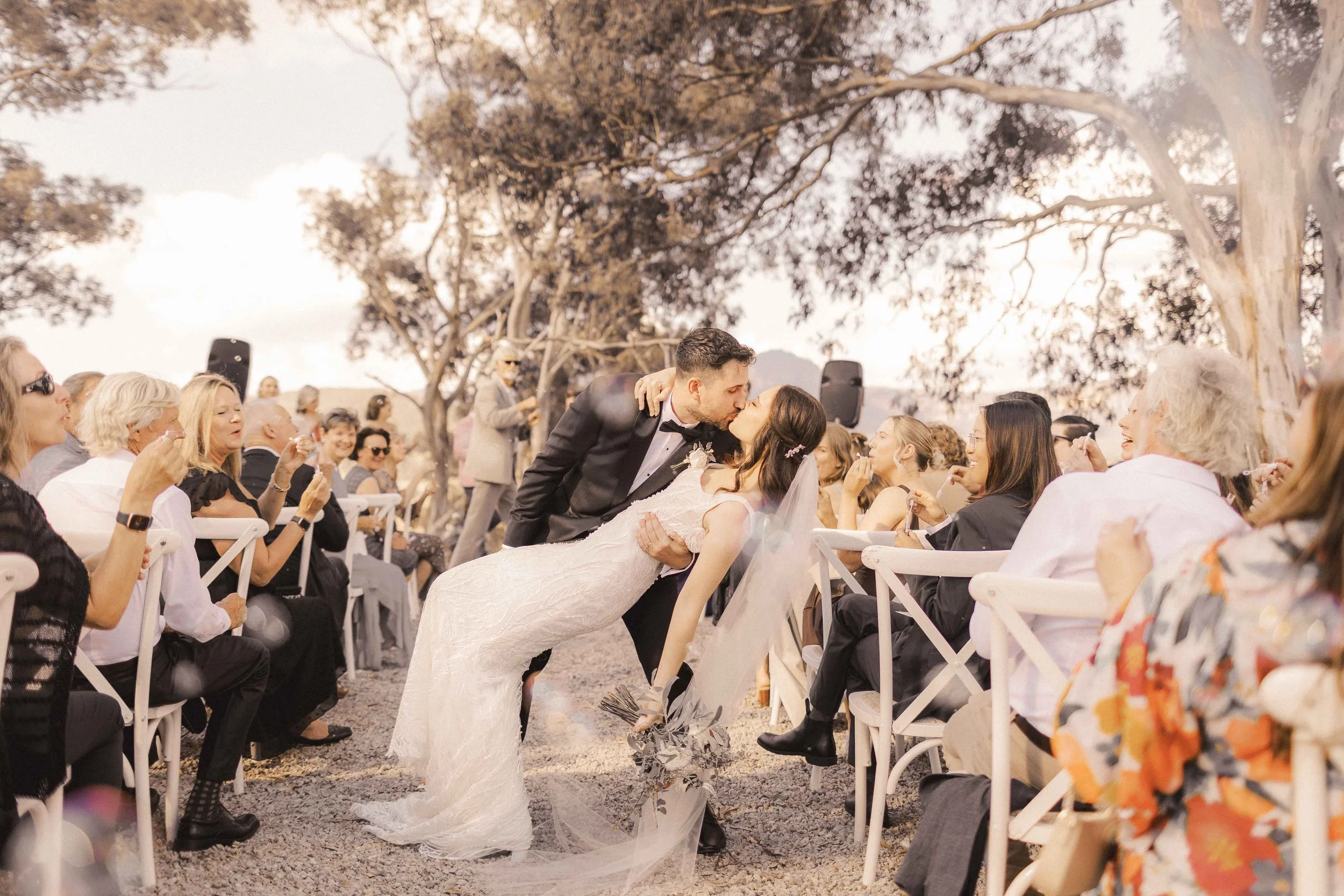 A bride and groom share a kiss at an outdoor wedding ceremony, surrounded by seated guests and large trees with overcast skies.