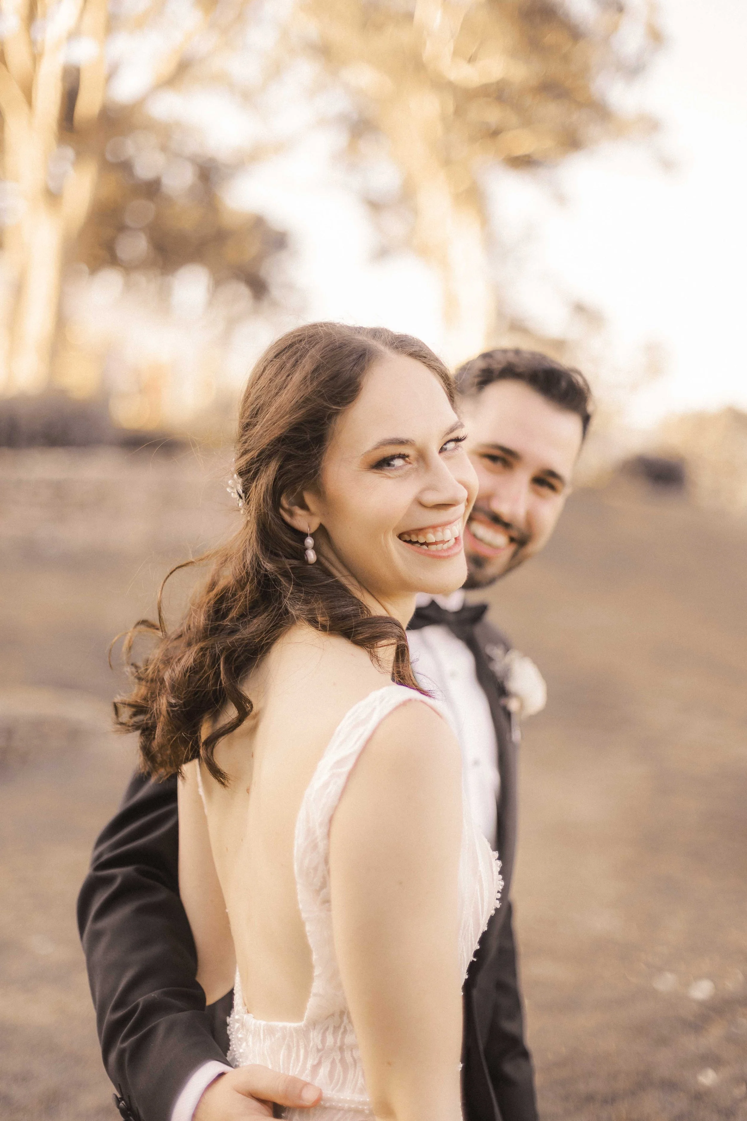 A bride and groom standing outdoors, smiling, with a blurred background of autumn trees and bright sunlight.