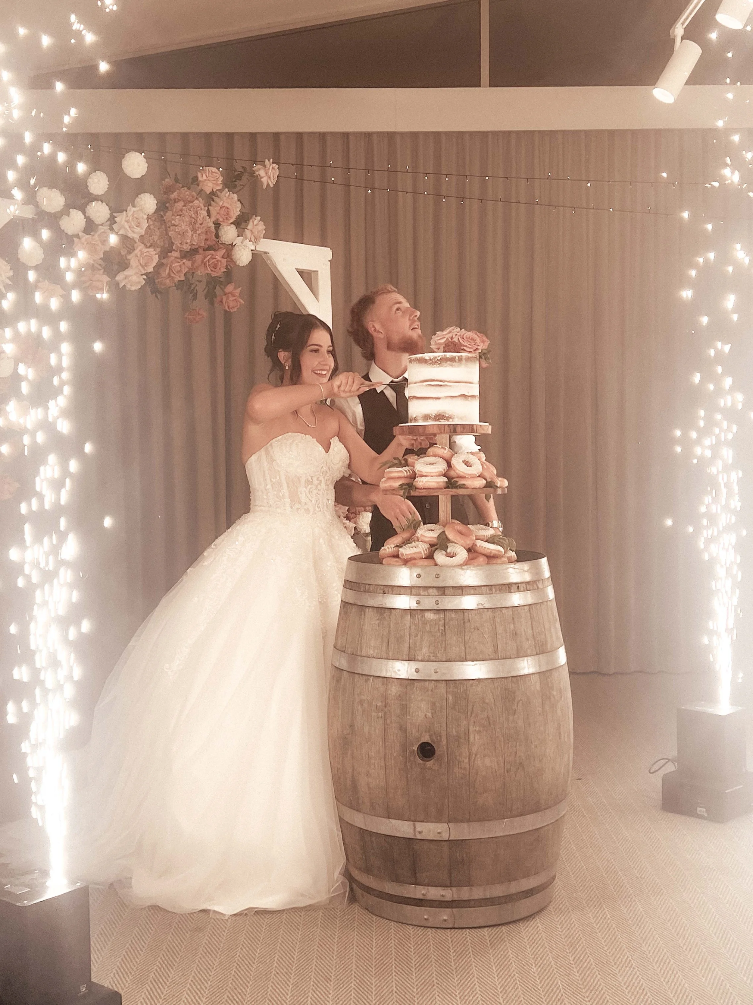 A bride and groom standing behind a wedding cake on a barrel, decorated with flowers, at their wedding reception with string lights and a floral backdrop.
