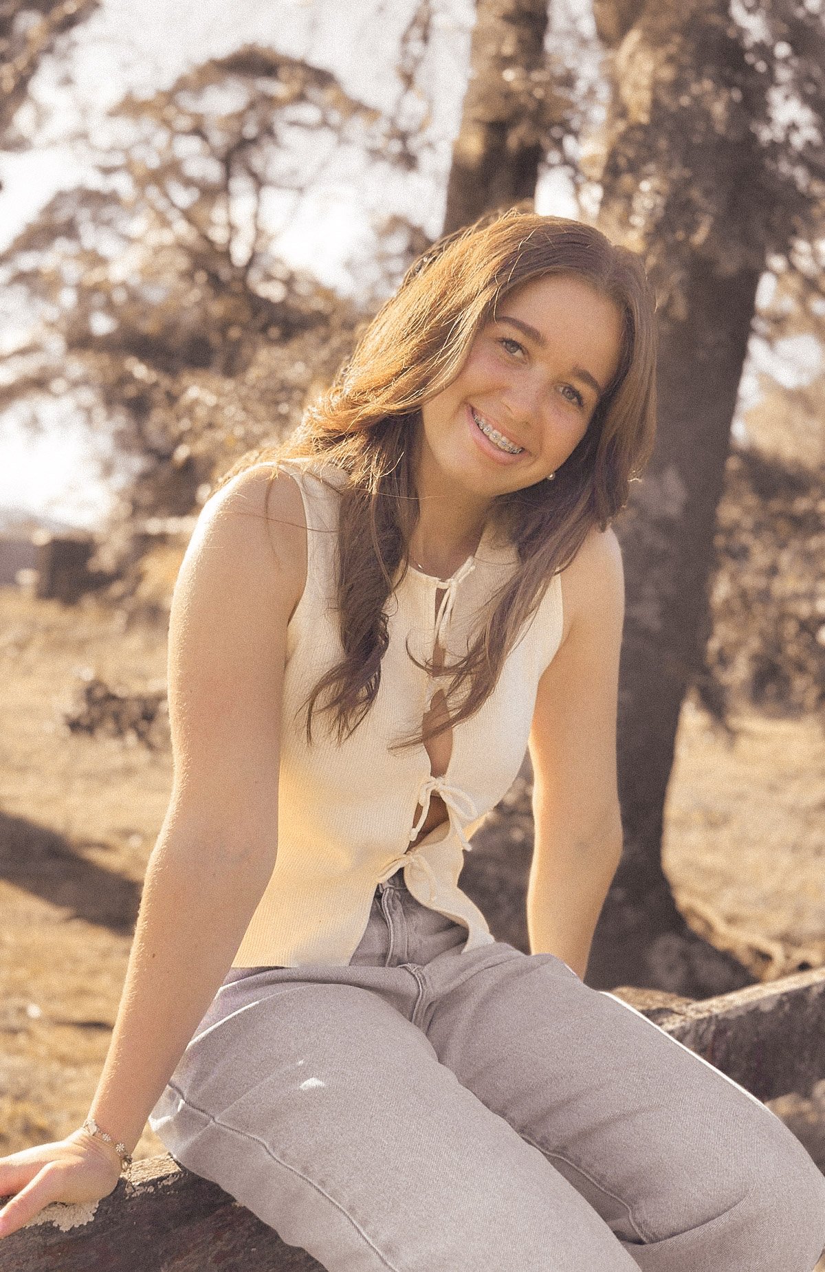 A young woman with long, wavy brown hair and braces smiling at the camera. She is sitting outdoors on a rock with bare trees and snow in the background, wearing a sleeveless off-white top and light-colored jeans.