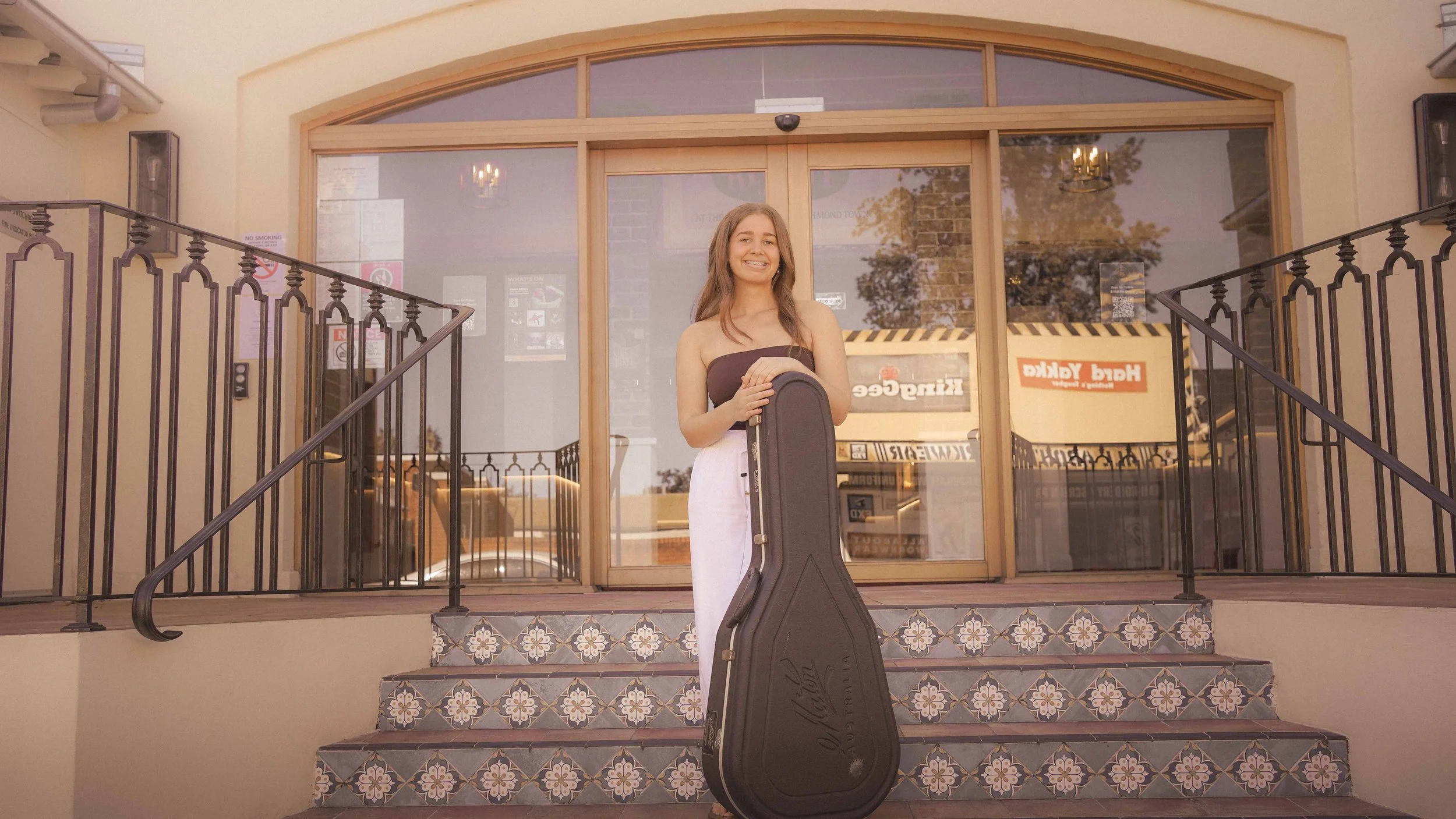 A young woman standing on front stairs outside a building, holding a guitar case, smiling, with a large window behind her reflecting trees and a vintage chandelier.