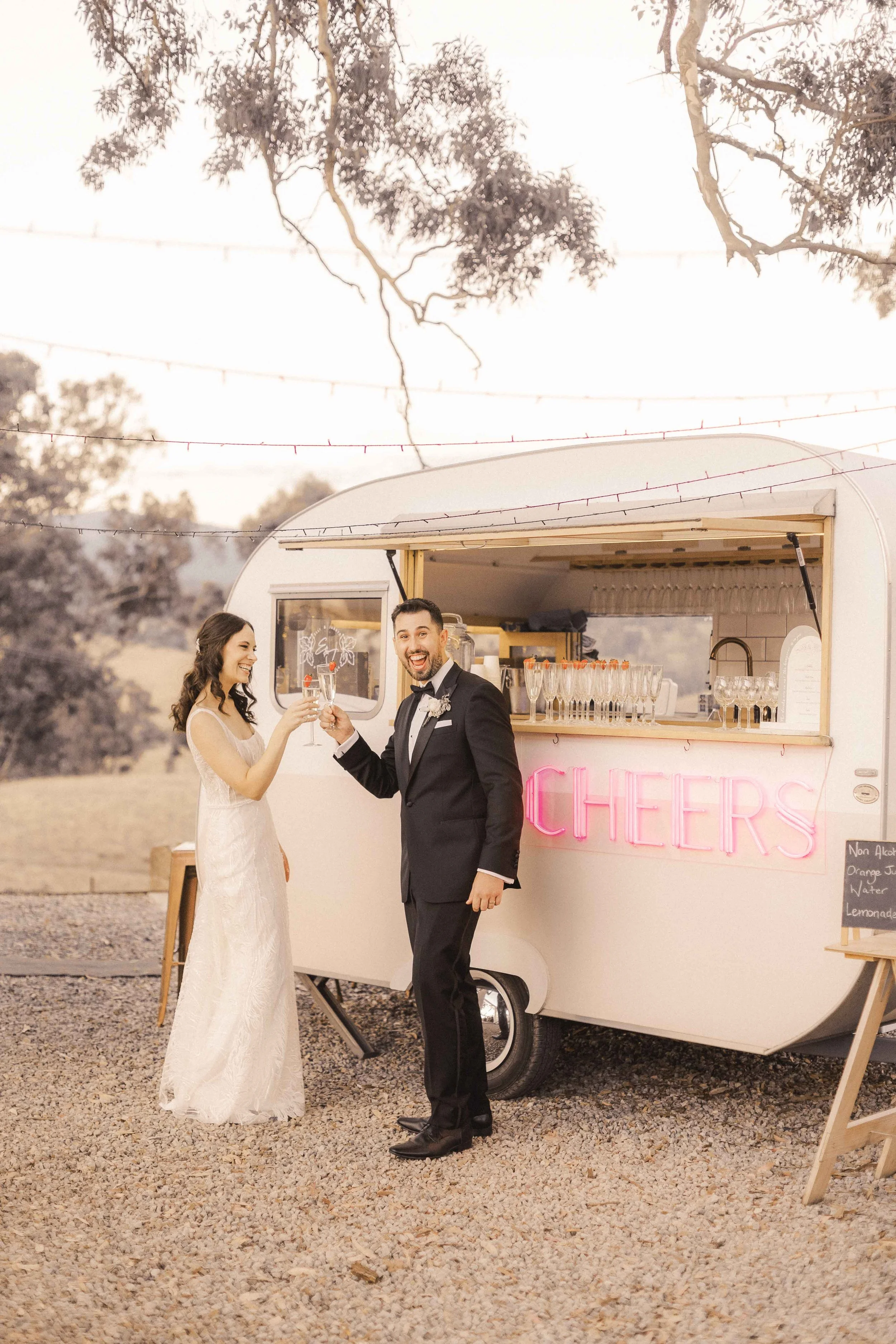 A bride and groom in front of a wedding food truck, holding champagne flutes, smiling and toasting each other at sunset outdoors.