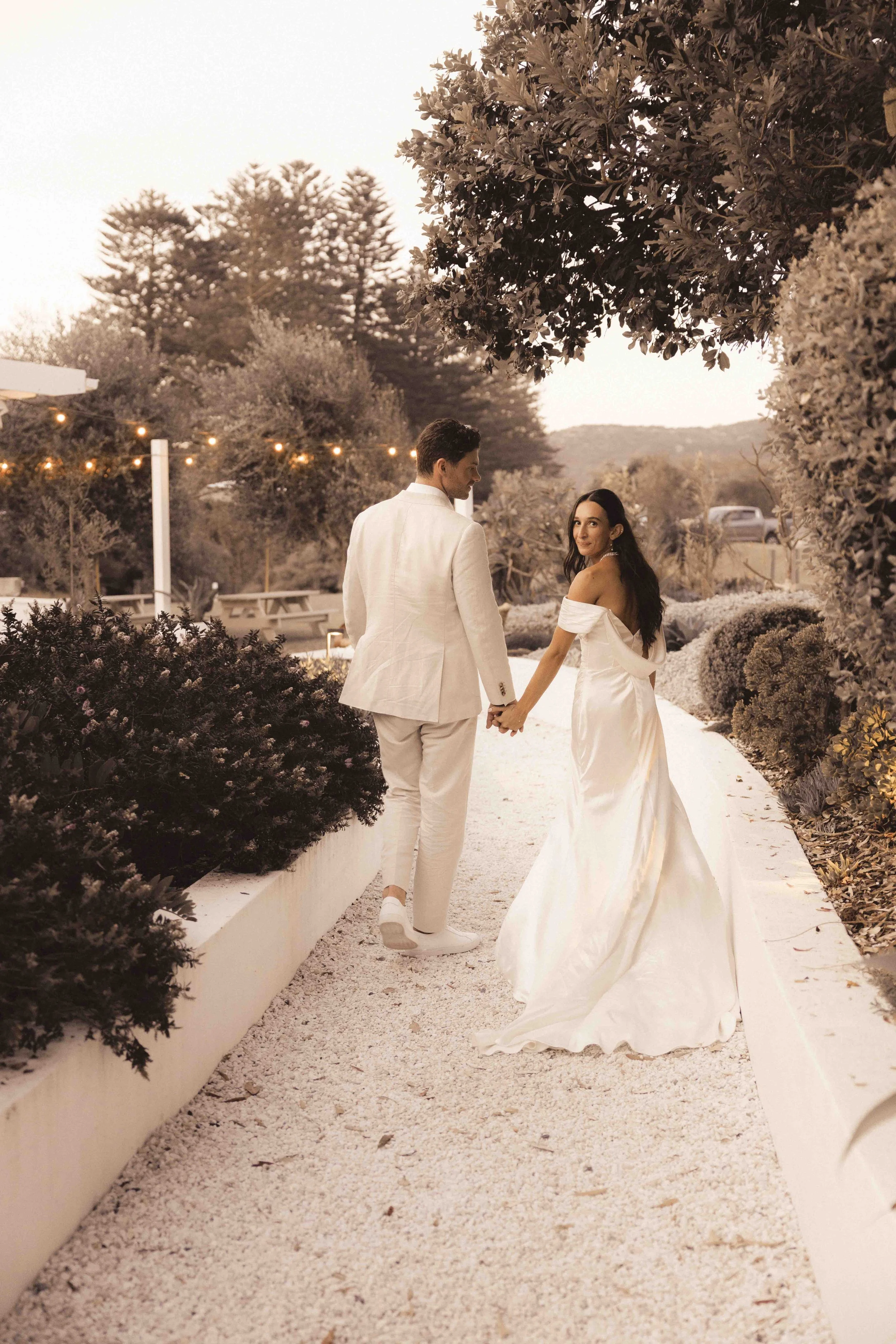 A couple dressed in white wedding attire holding hands and facing each other, standing outdoors on a white gravel pathway surrounded by bushes and trees, with string lights hanging in the background during dusk or early evening.
