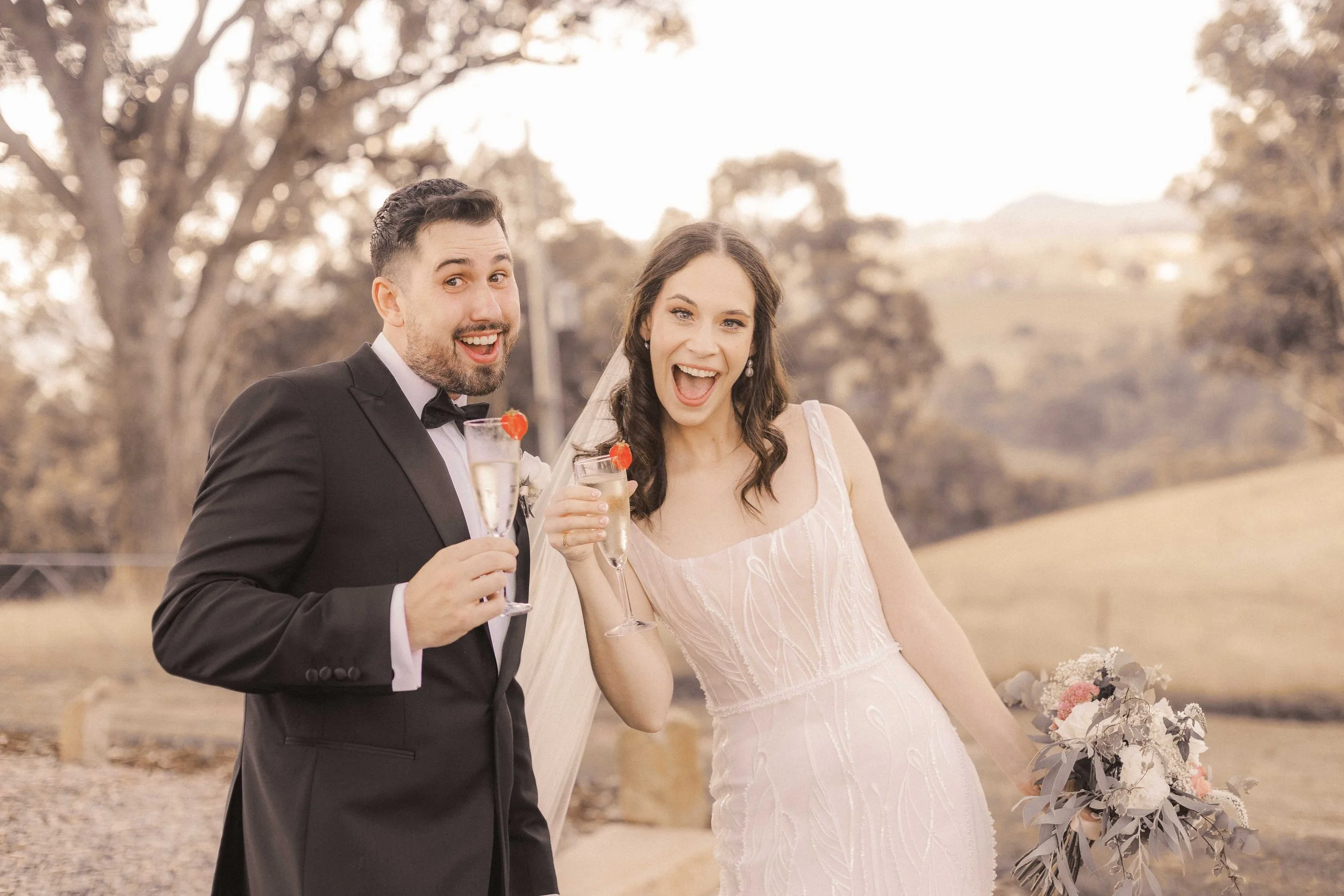 A bride and groom celebrating outdoors, holding glasses of champagne with strawberries, smiling happily. The groom wears a black tuxedo, and the bride wears a white wedding dress, holding a bouquet of flowers. The background includes trees and a soft