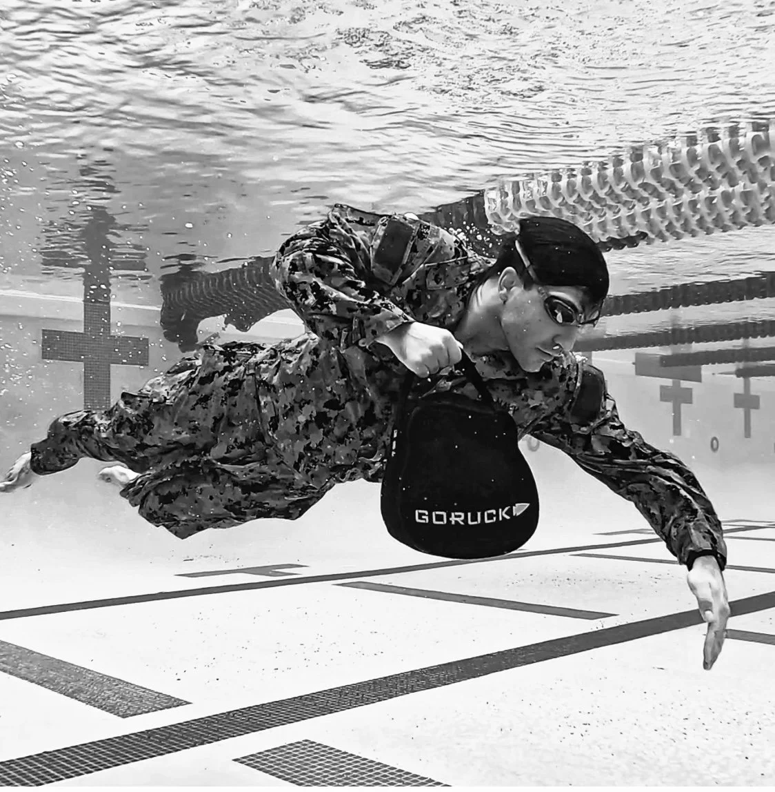 Soldier in military uniform swimming underwater in a swimming pool, holding a weighted bag labeled 'GORUCK'.