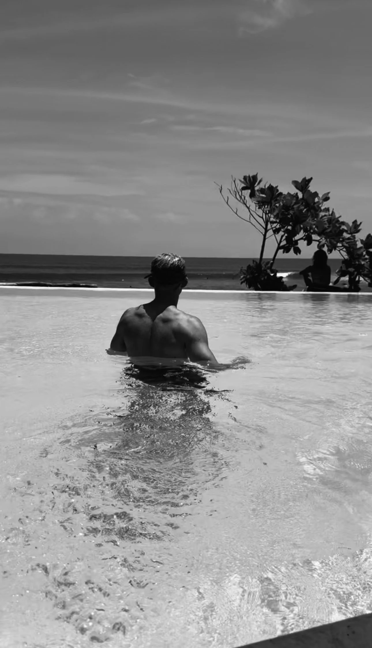 A man with sunglasses sitting in a swimming pool facing the ocean, with a person sitting on the deck and a tree in the background.