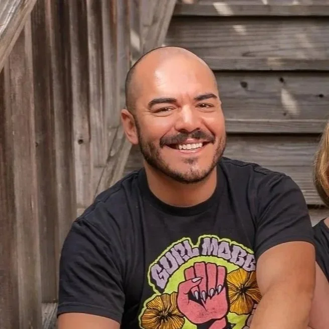 A smiling man with a beard and shaved head, sitting outdoors on wooden stairs, wearing a black t-shirt with a graphic design and black shorts.