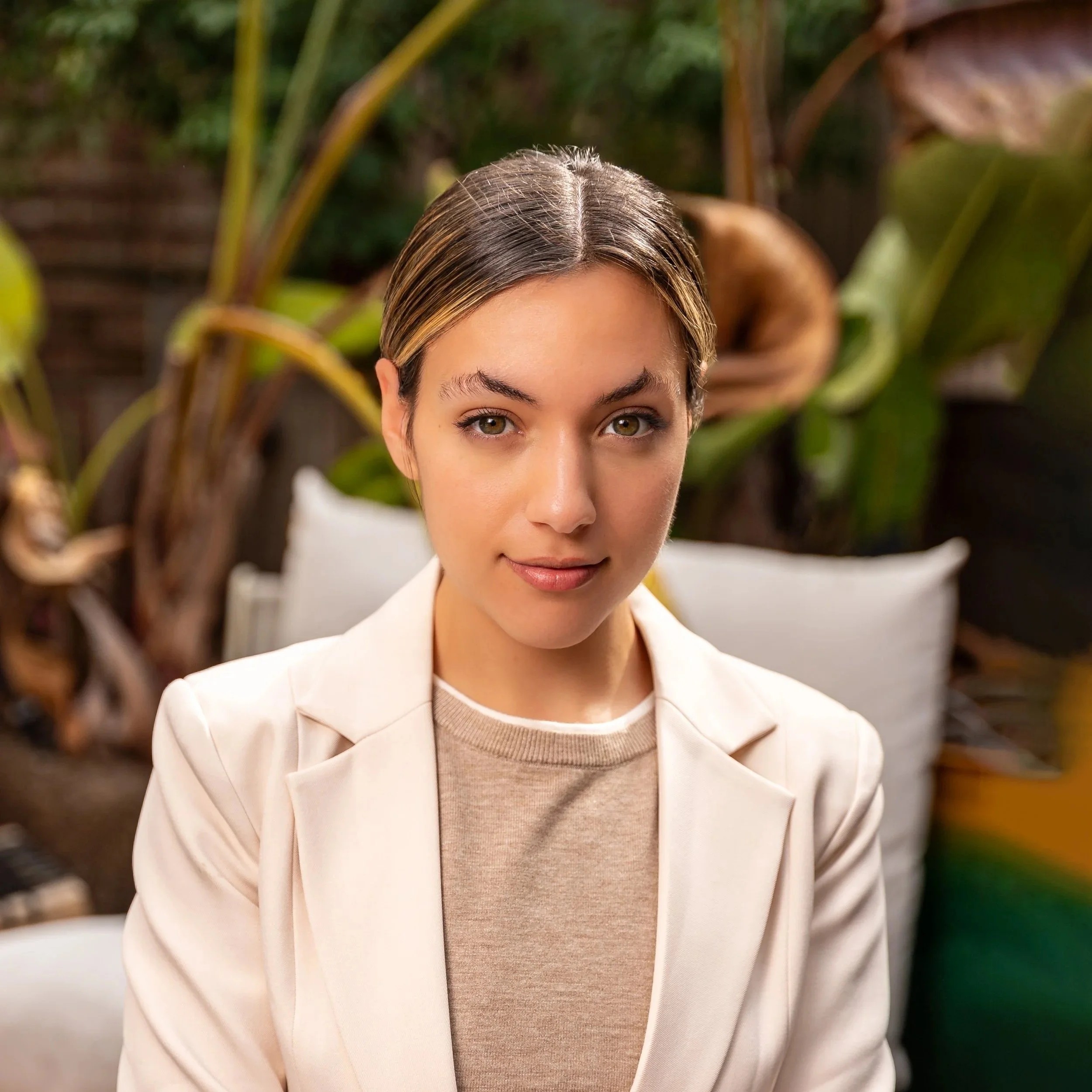 A young woman in a beige blazer and light brown shirt sitting outdoors with green plants in the background.